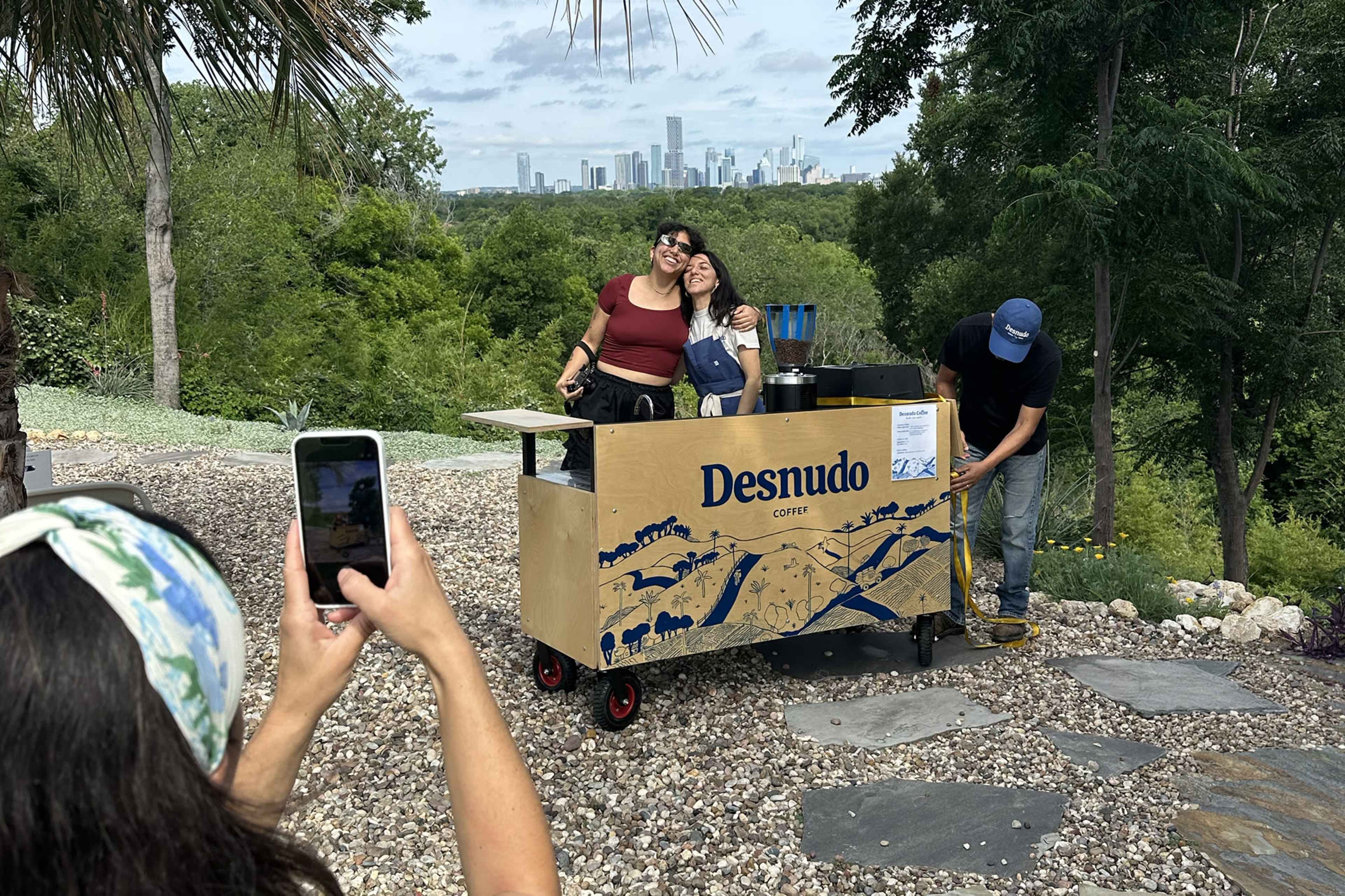 Two women smile behind a coffee cart labeled "Desnudo," while a man prepares drinks in a scenic outdoor setting with a city skyline in the background.