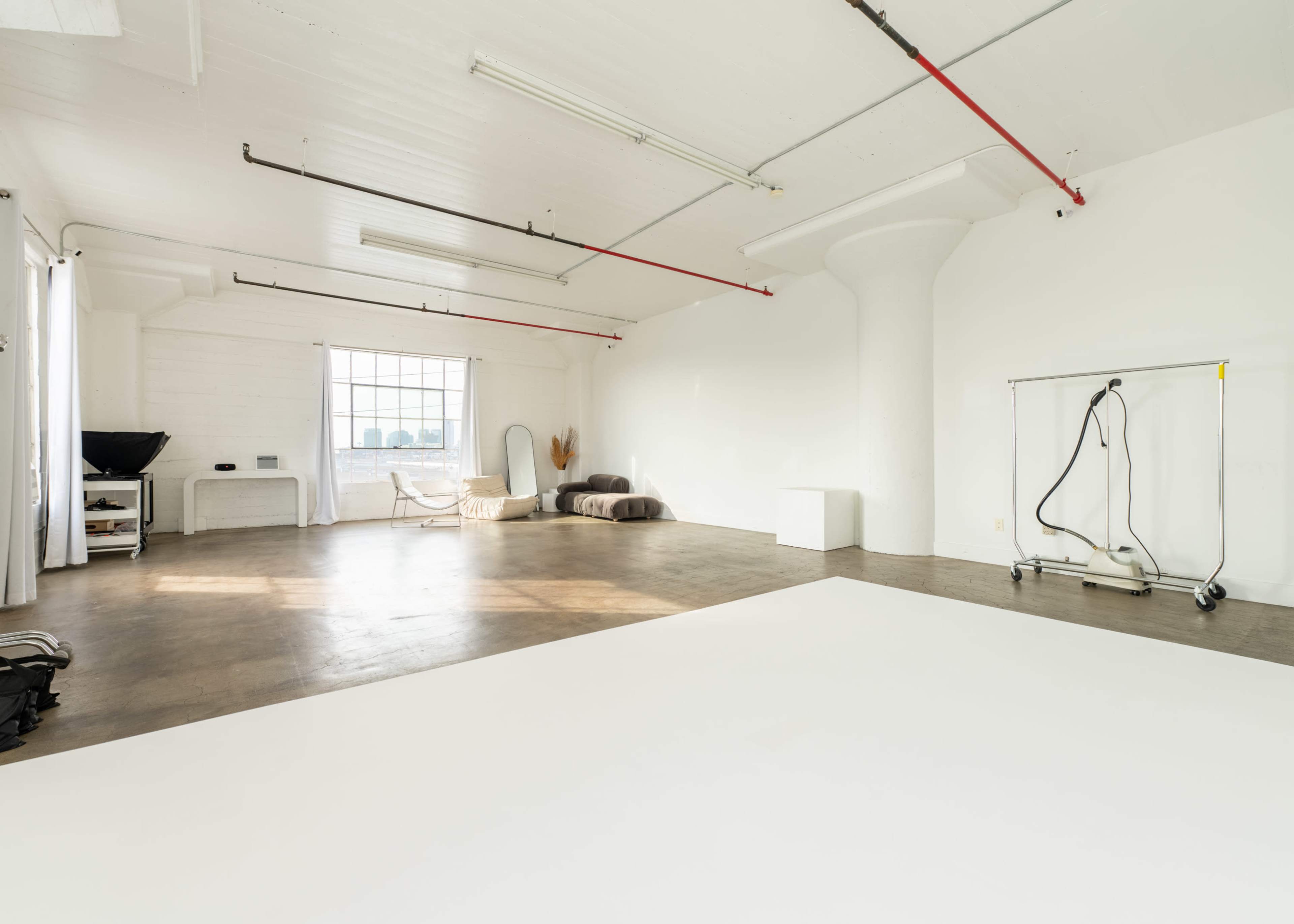 The image shows a spacious, well-lit studio with white walls and a concrete floor, featuring a large window, minimal furniture, and a rolling clothing rack.