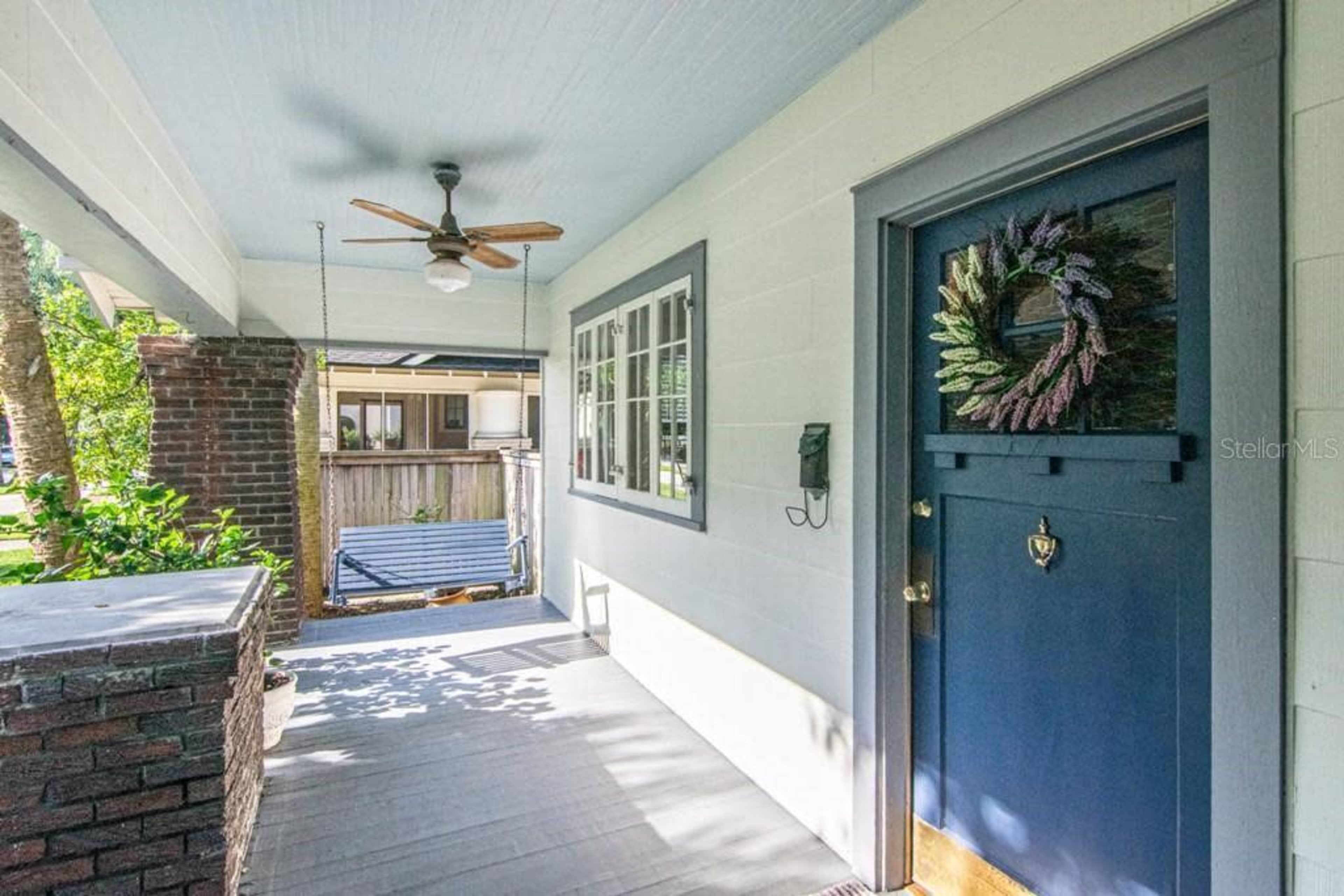 The image shows a porch area with a navy blue front door adorned with a wreath, a ceiling fan, and a swing behind a brick column.