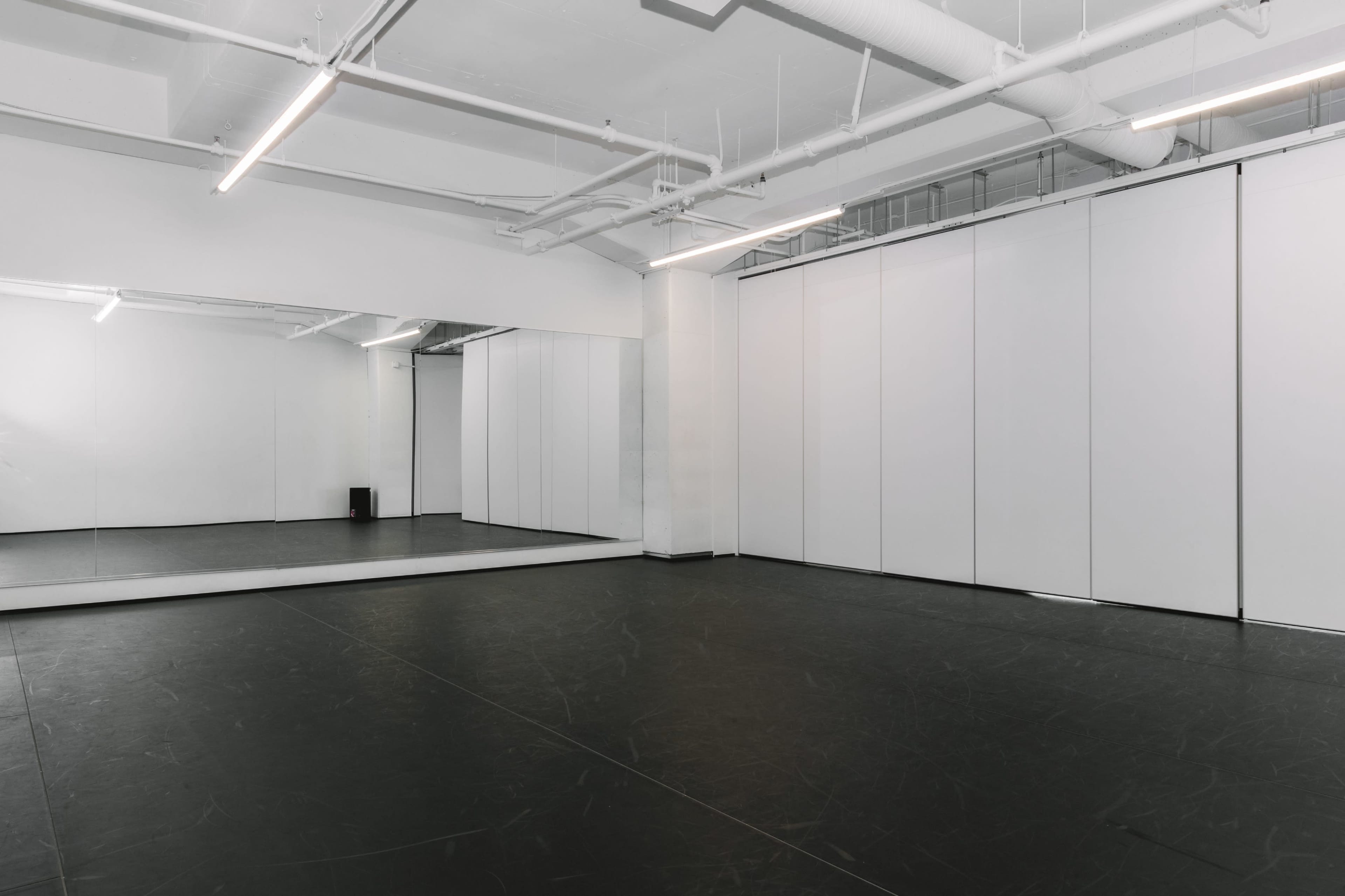 The image depicts a spacious, empty dance studio with a large mirror on one wall and smooth black flooring.