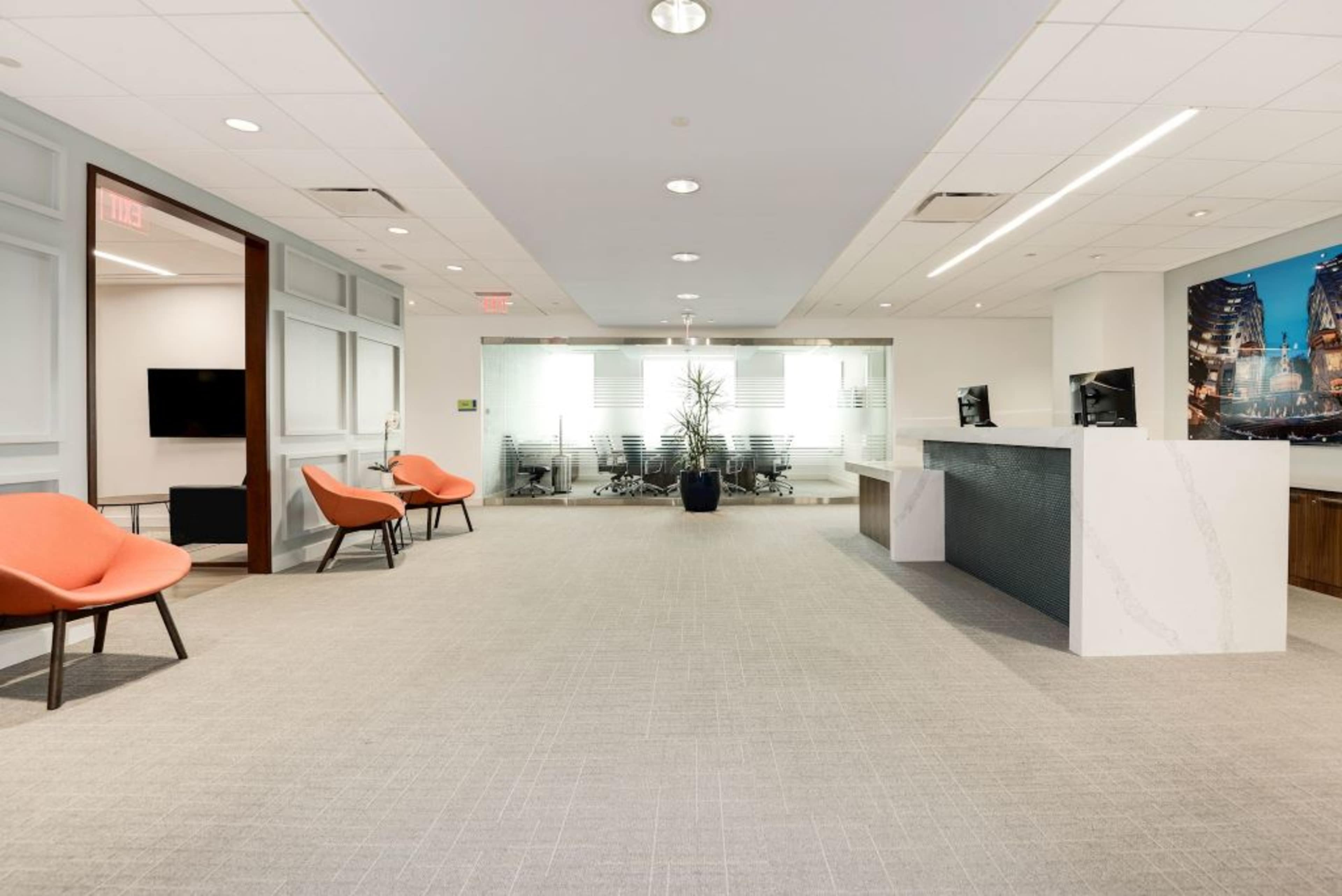 A modern office reception area featuring two orange chairs, a reception desk, and a glass-enclosed conference room.