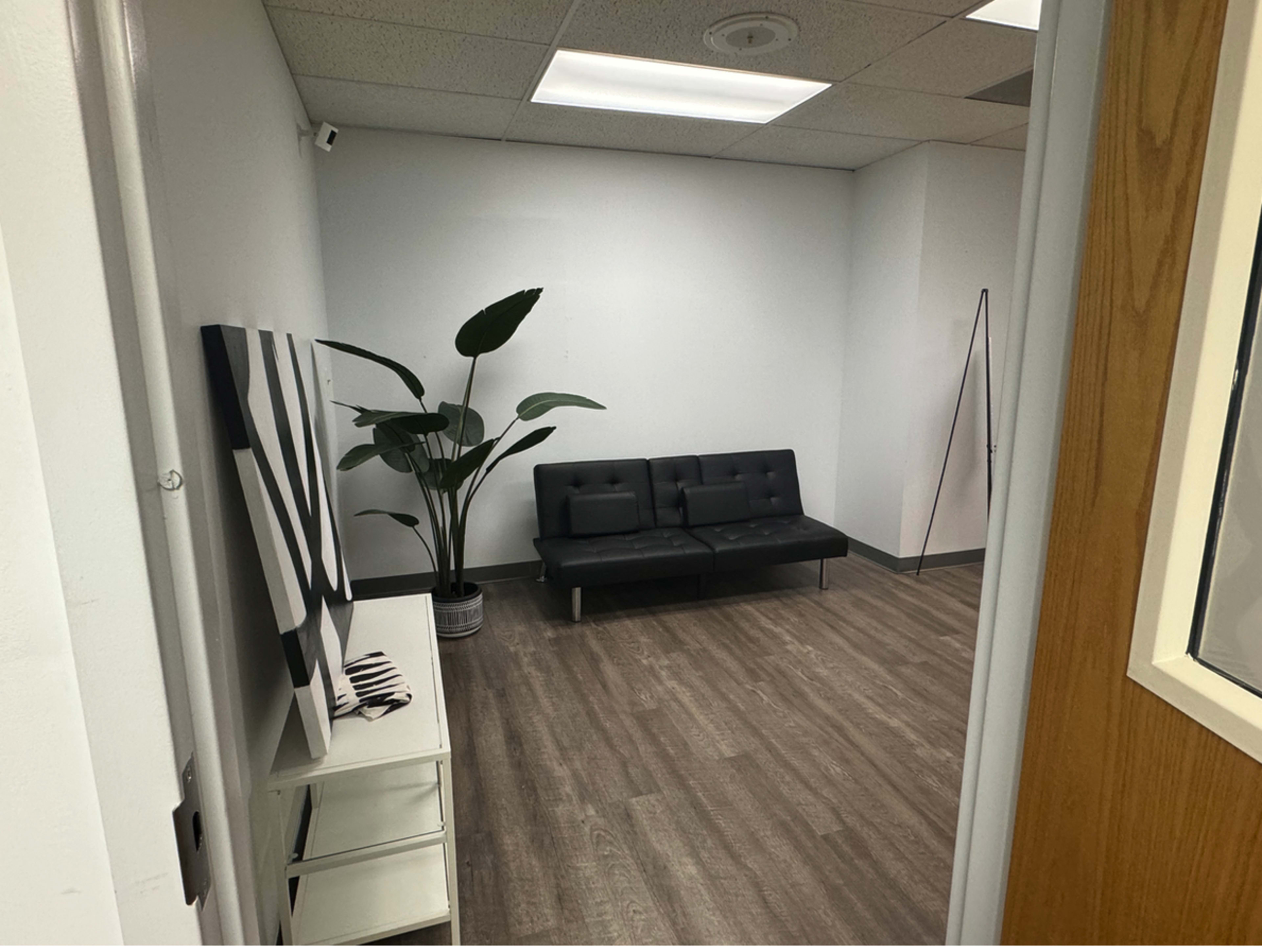 The image shows a minimalist office room with a black sofa, a potted plant, and a white cabinet with decorative items.