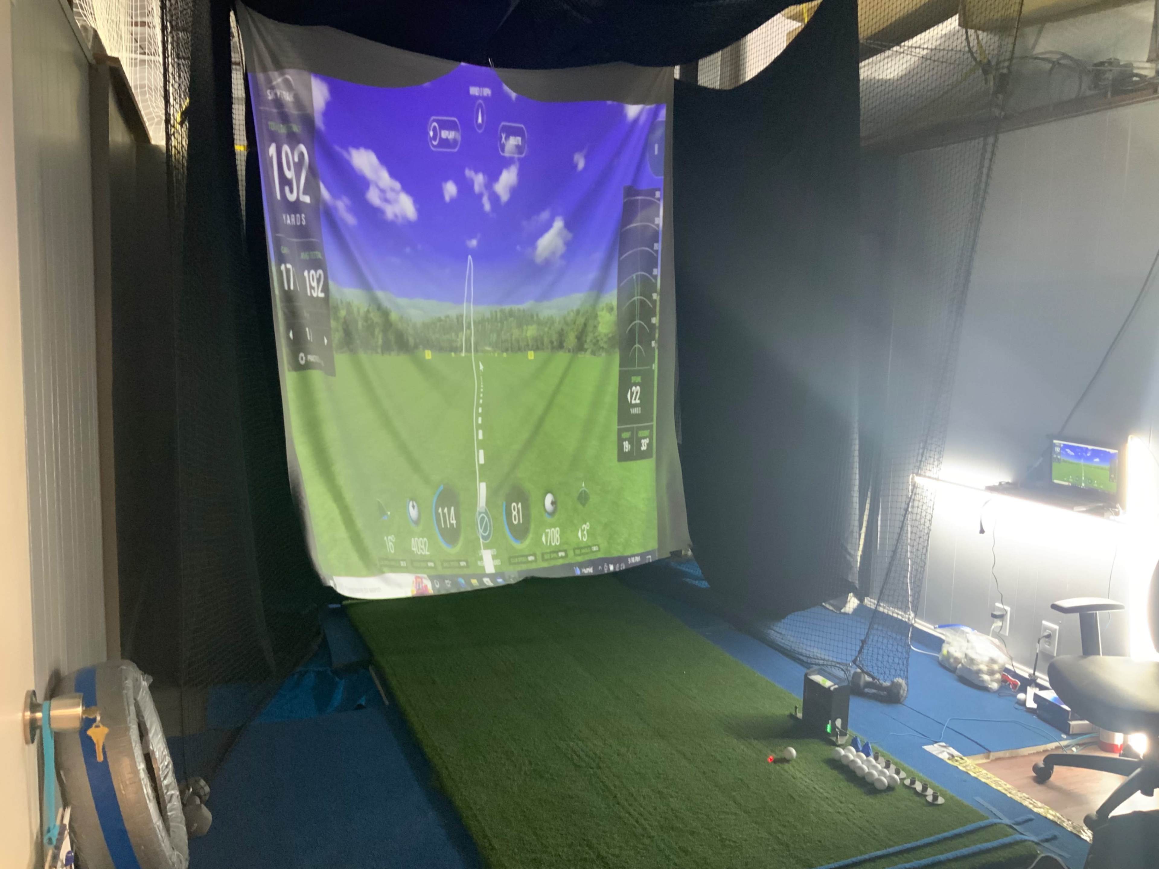 The image shows an indoor golf simulator setup with a large screen displaying golf course graphics and a synthetic turf mat.