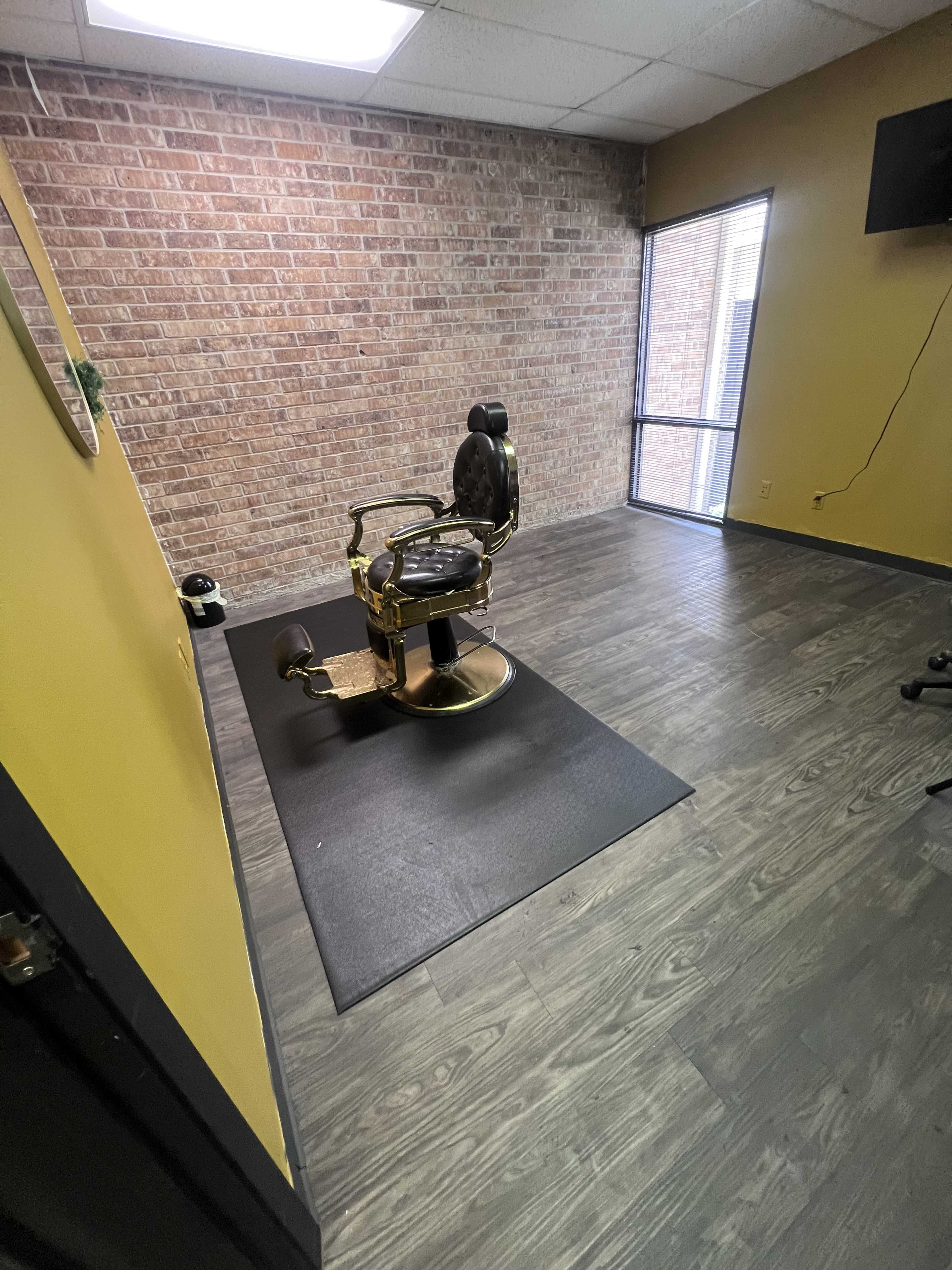 The image shows a barber chair positioned on a black mat in a room with a brick wall and yellow-painted walls.