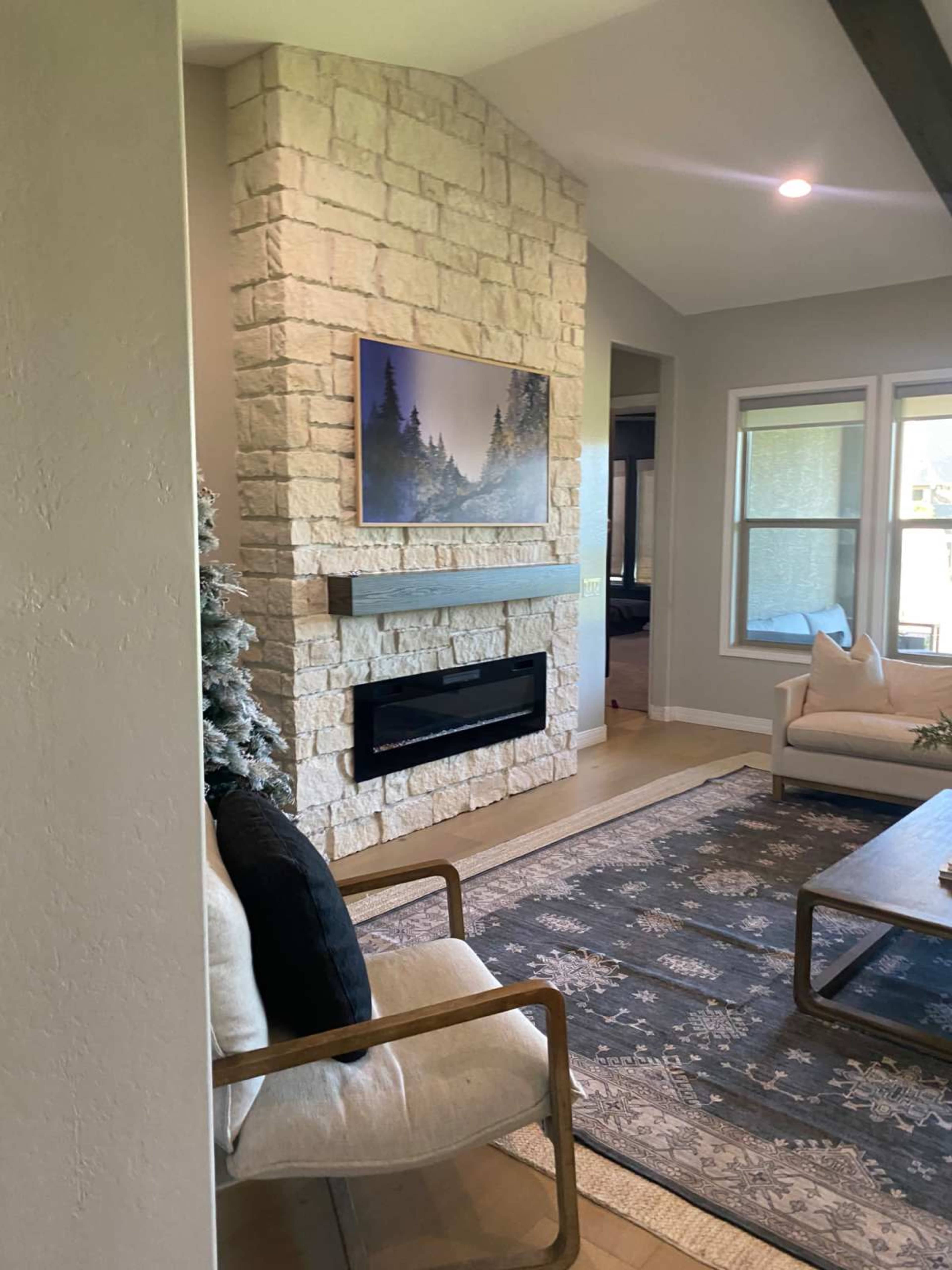 The image shows a modern living room with a stone fireplace, a large artwork above it, and light-colored furniture arranged around a patterned area rug.