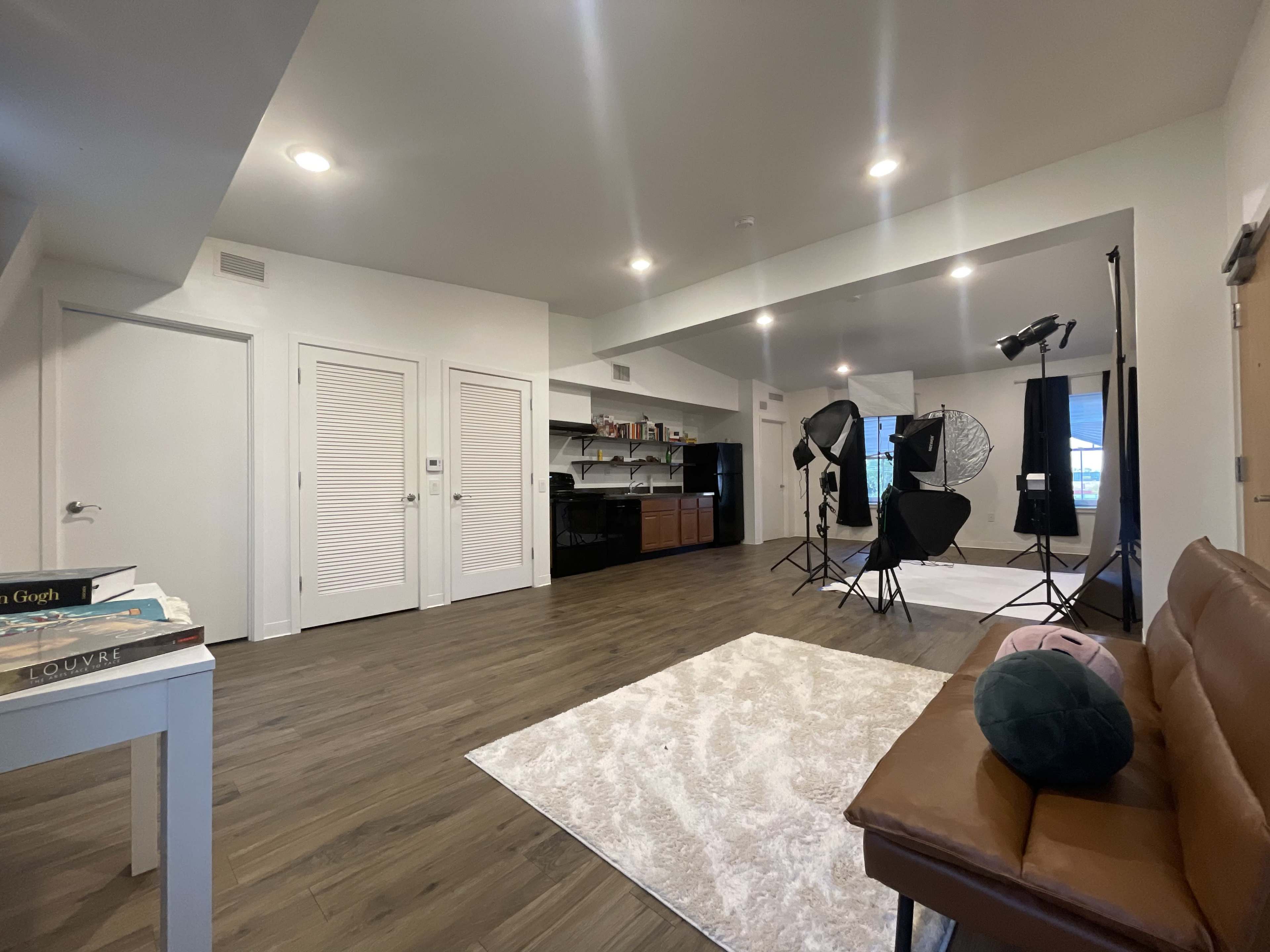 A spacious interior living area with a brown sofa, a white rug, kitchen cabinets, and photography equipment set up in the background.