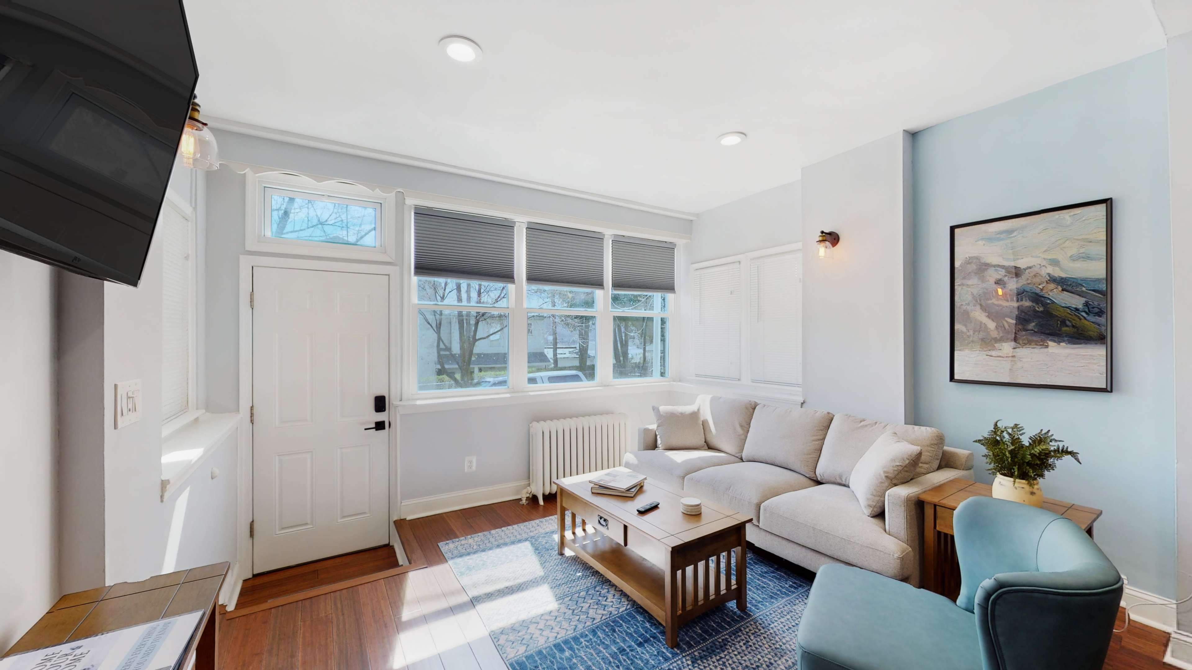 A bright living room with a light-colored sofa, a wooden coffee table, and a framed painting on the wall near a large window.