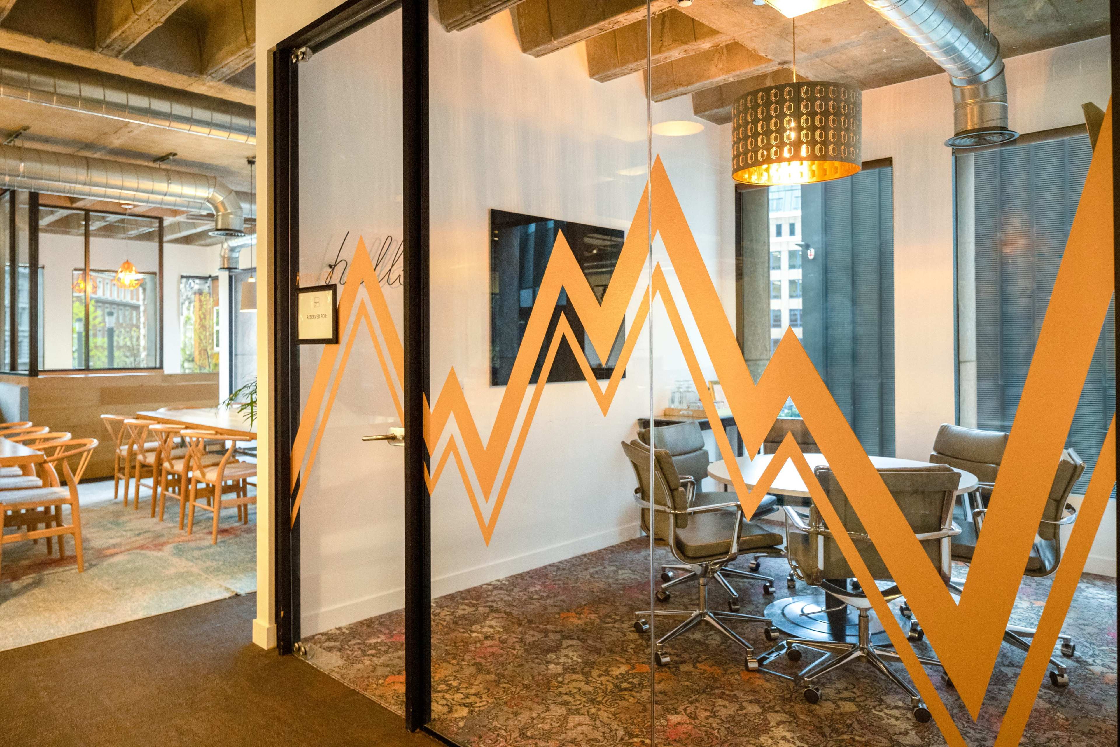 The image shows a modern office space with a glass door featuring an orange zigzag design, and a meeting area with chairs and a table inside.