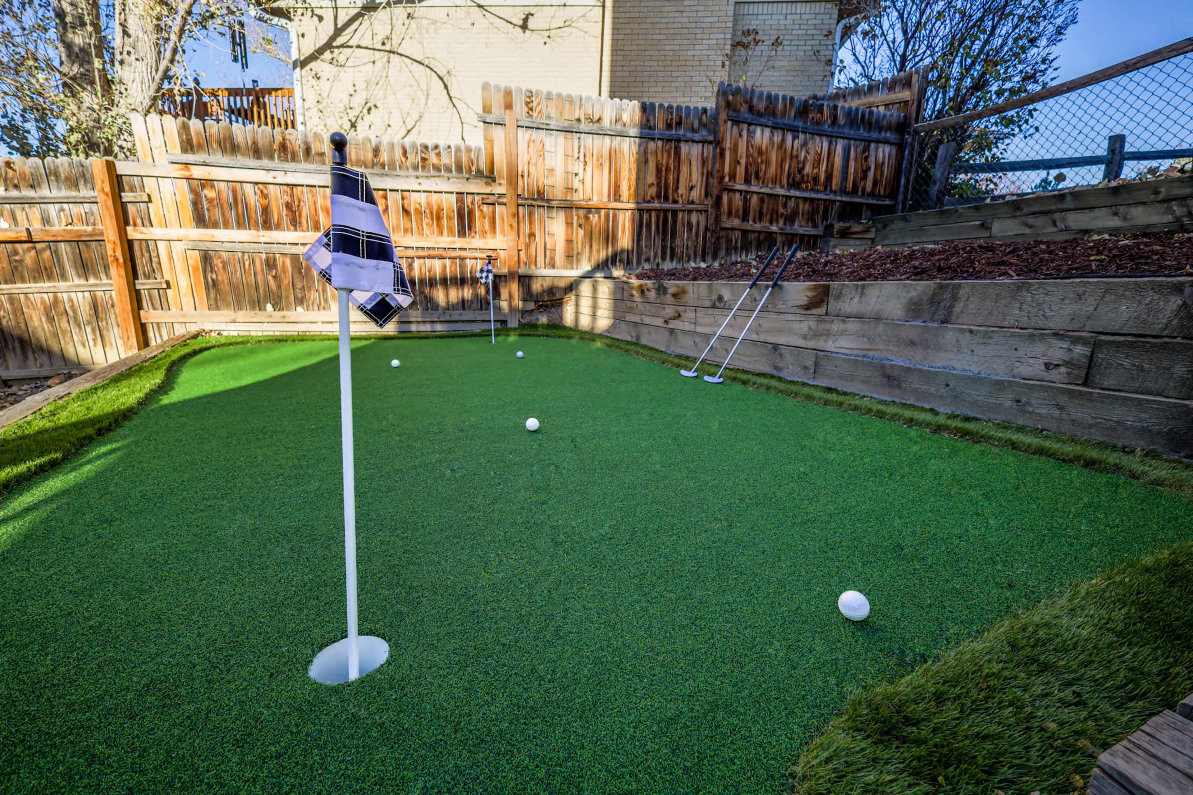 A small putting green in a backyard, featuring a flag in a hole and several golf balls on the artificial grass surface.