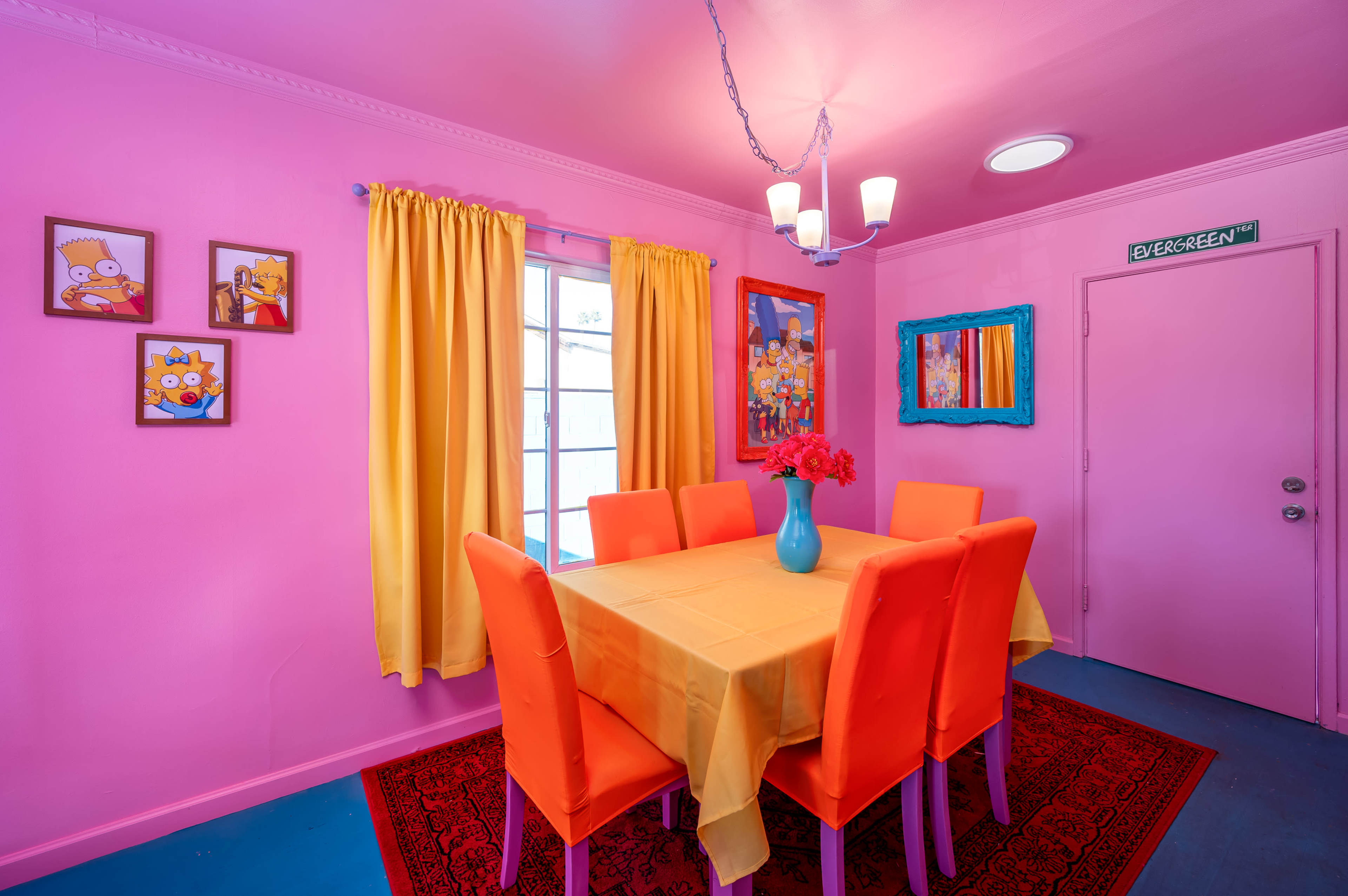 The dining area features a pink wall, a table covered with a yellow tablecloth, and bright orange chairs, surrounded by framed artwork and yellow curtains.