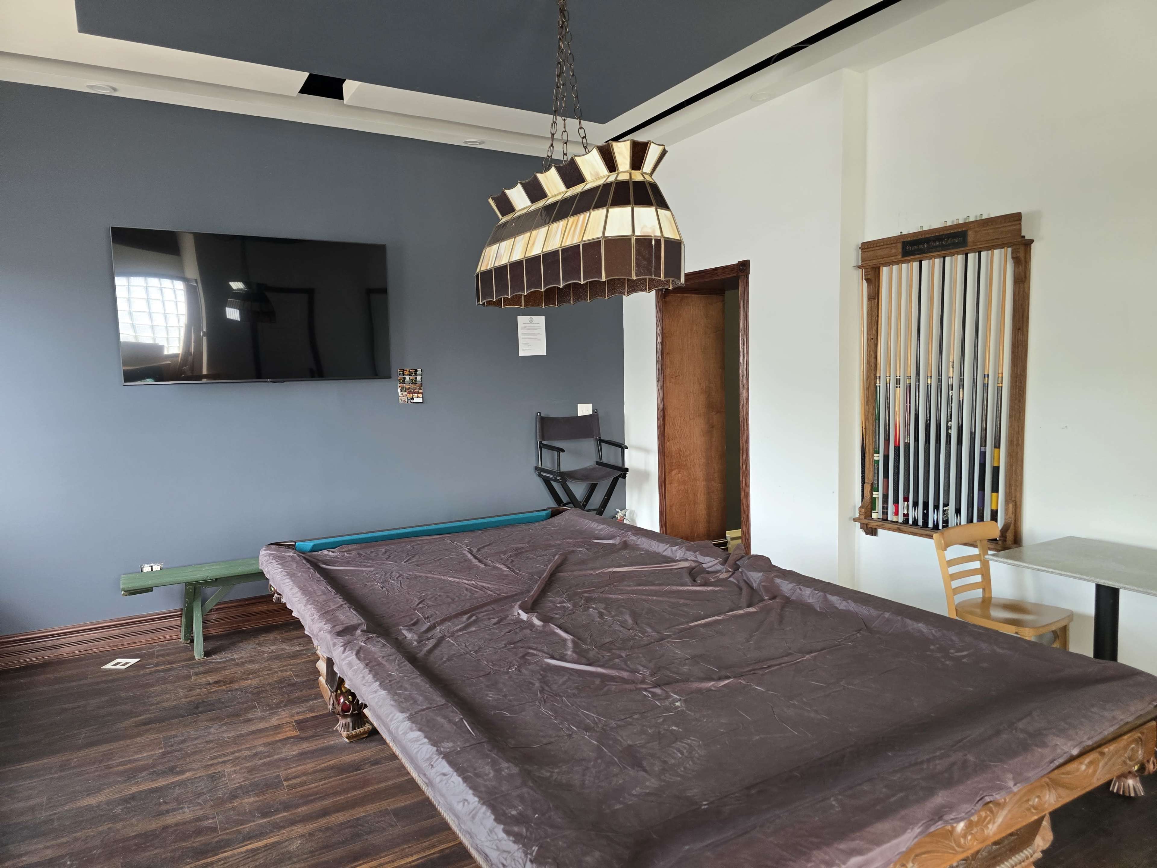 The image shows a room with a billiards table covered by a brown cloth, a wall-mounted television, and a rack for cues beside a small table and a chair.