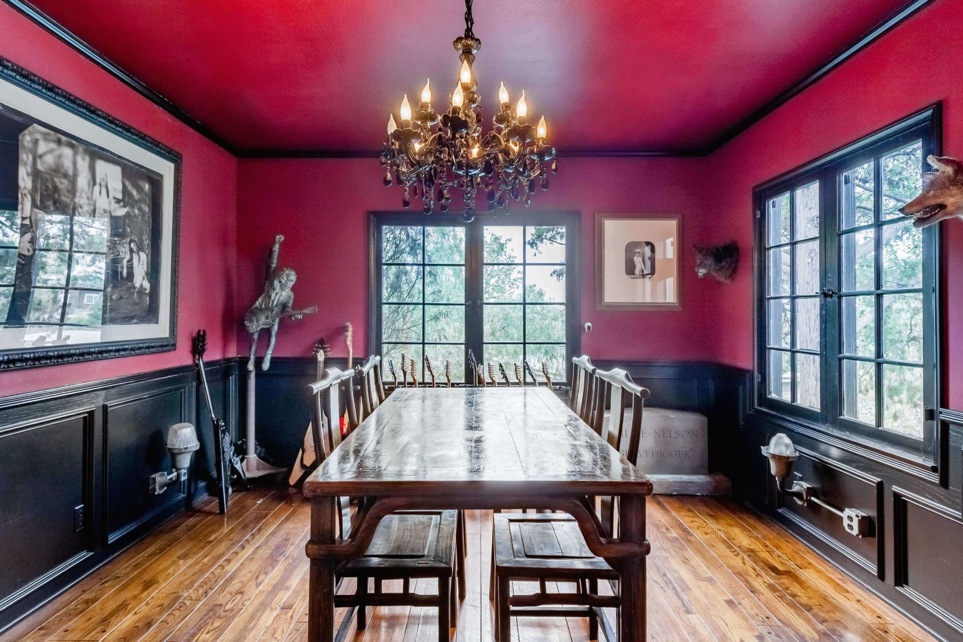 The dining room features a large wooden table surrounded by chairs, illuminated by a chandelier, with red walls and large windows overlooking a natural landscape.