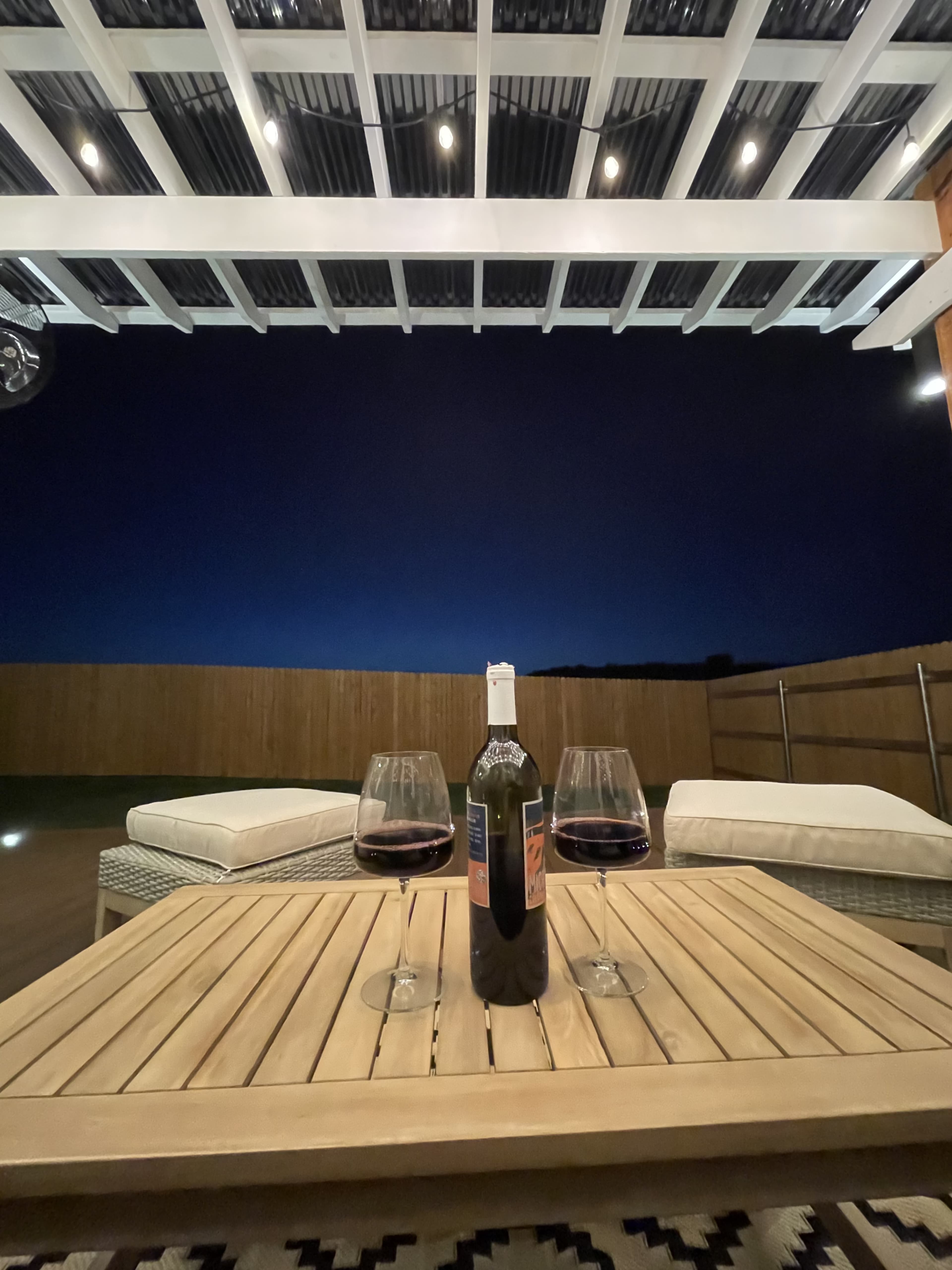 A table with two glasses of red wine and a bottle sits on a patio under a pergola, with a dark blue sky in the background.