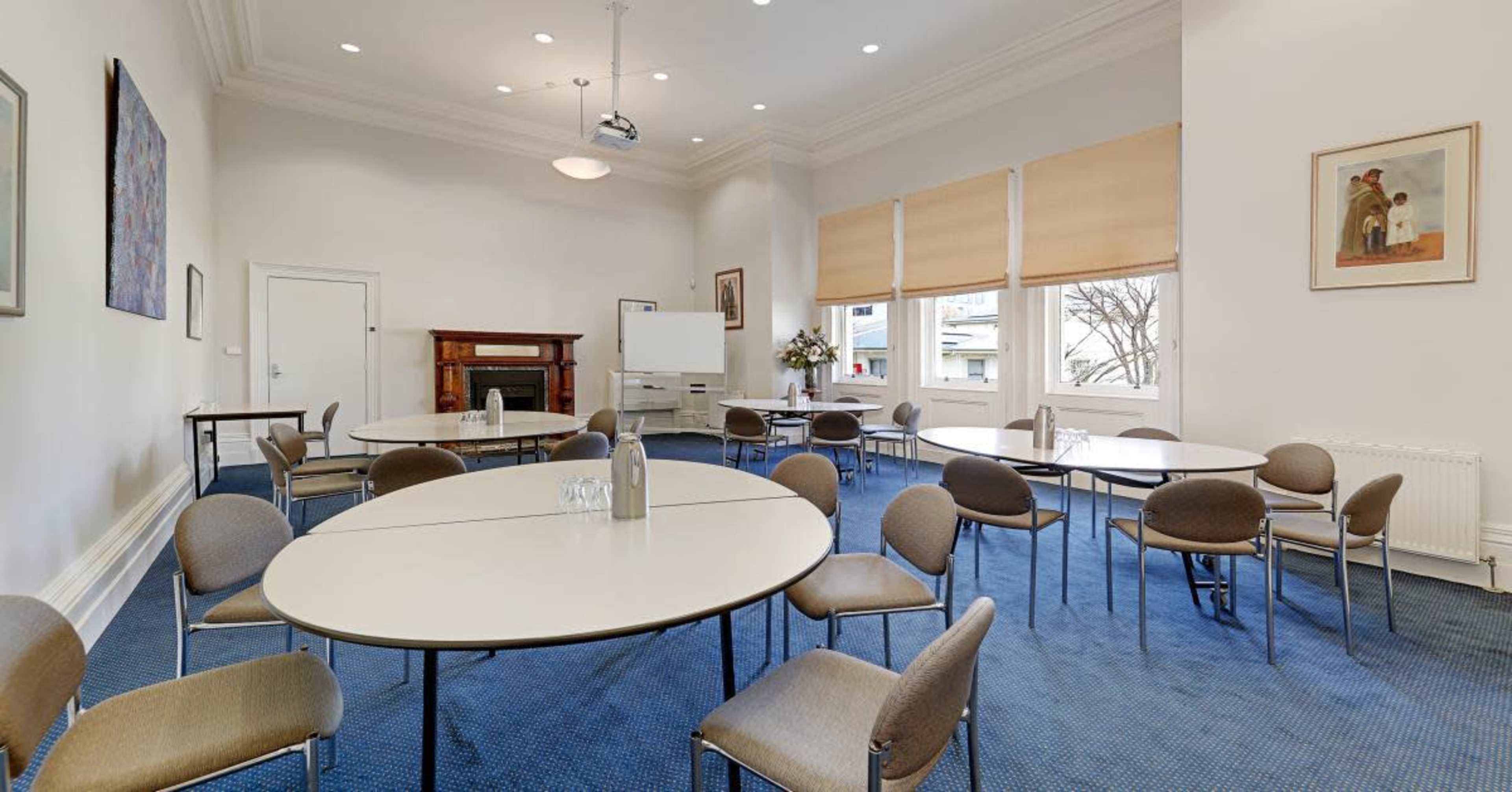 The image shows a well-lit meeting room with several round tables and chairs arranged neatly, along with a whiteboard and a decorative fireplace.