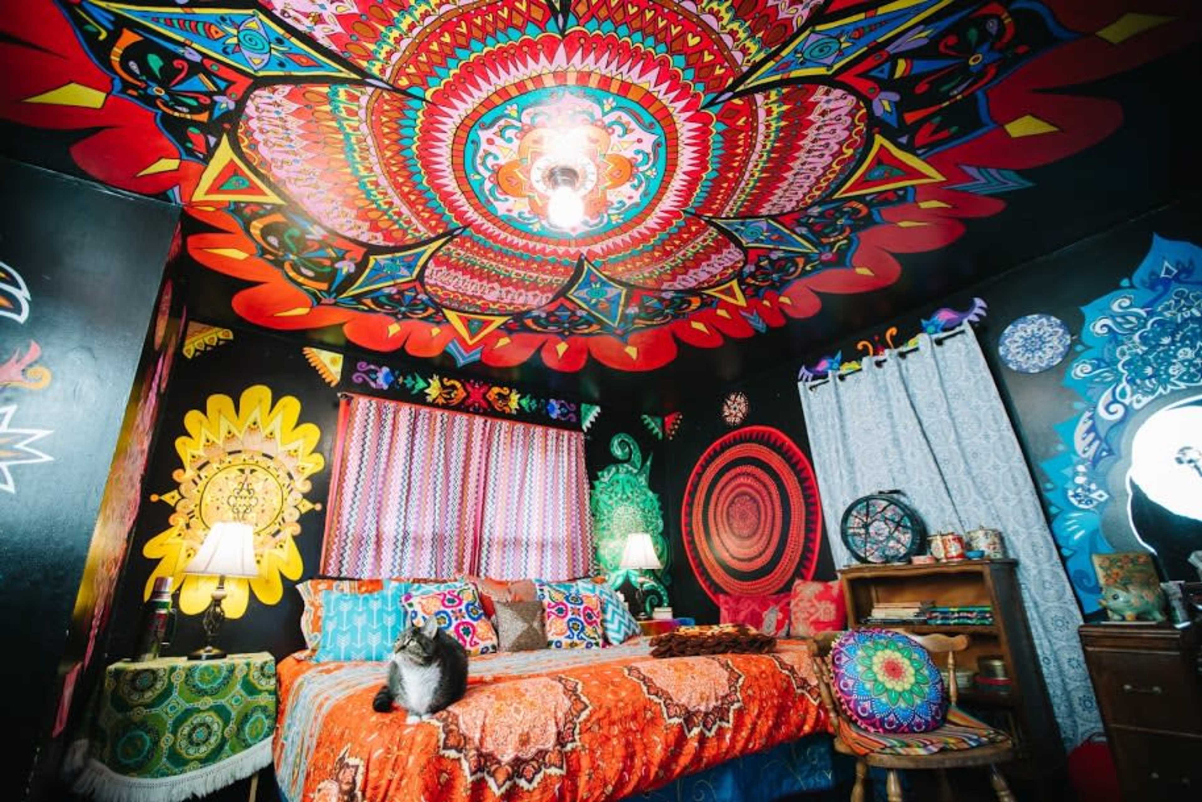 A vibrantly colored bedroom features a detailed, multicolored ceiling design and bold wall art, with a patterned bedspread and a cat sitting on the bed.