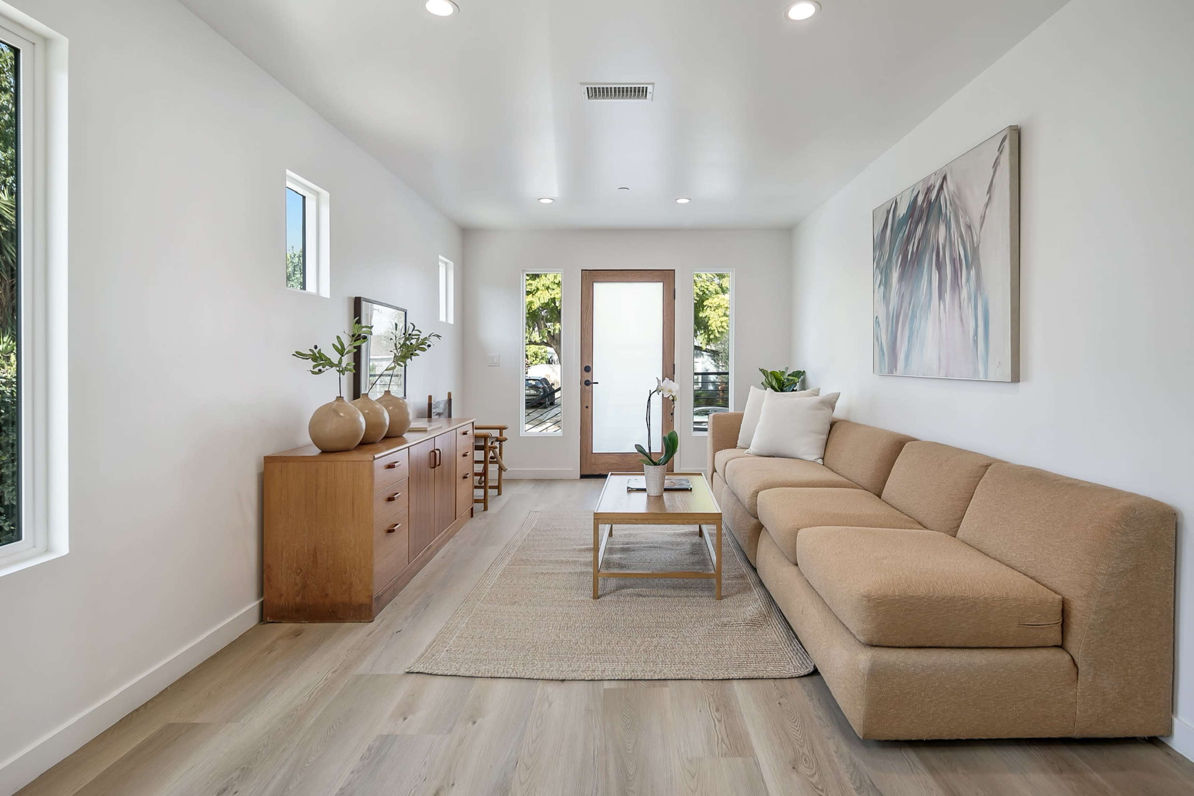 A modern living room features a beige sectional sofa, a wooden sideboard, and a coffee table on a light-colored rug, with large windows allowing natural light to enter.