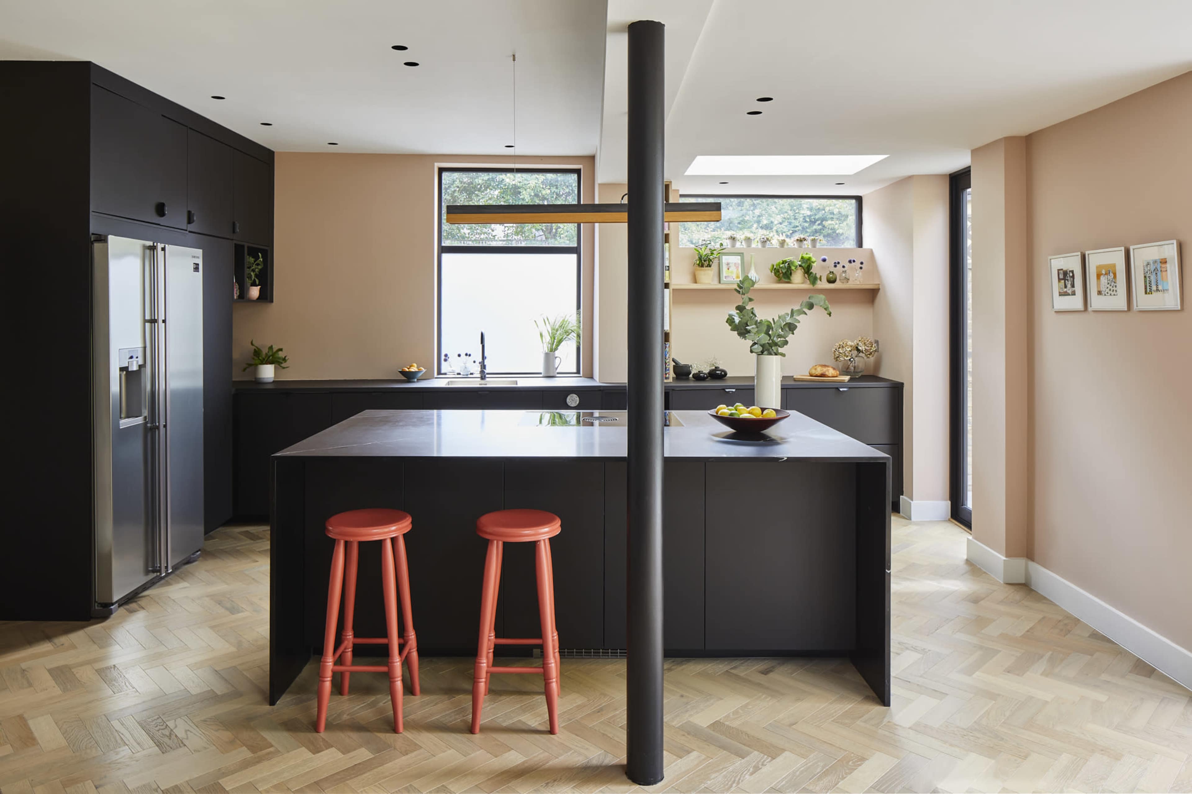 A modern kitchen features black cabinetry, a central island with orange bar stools, and a window overlooking greenery.