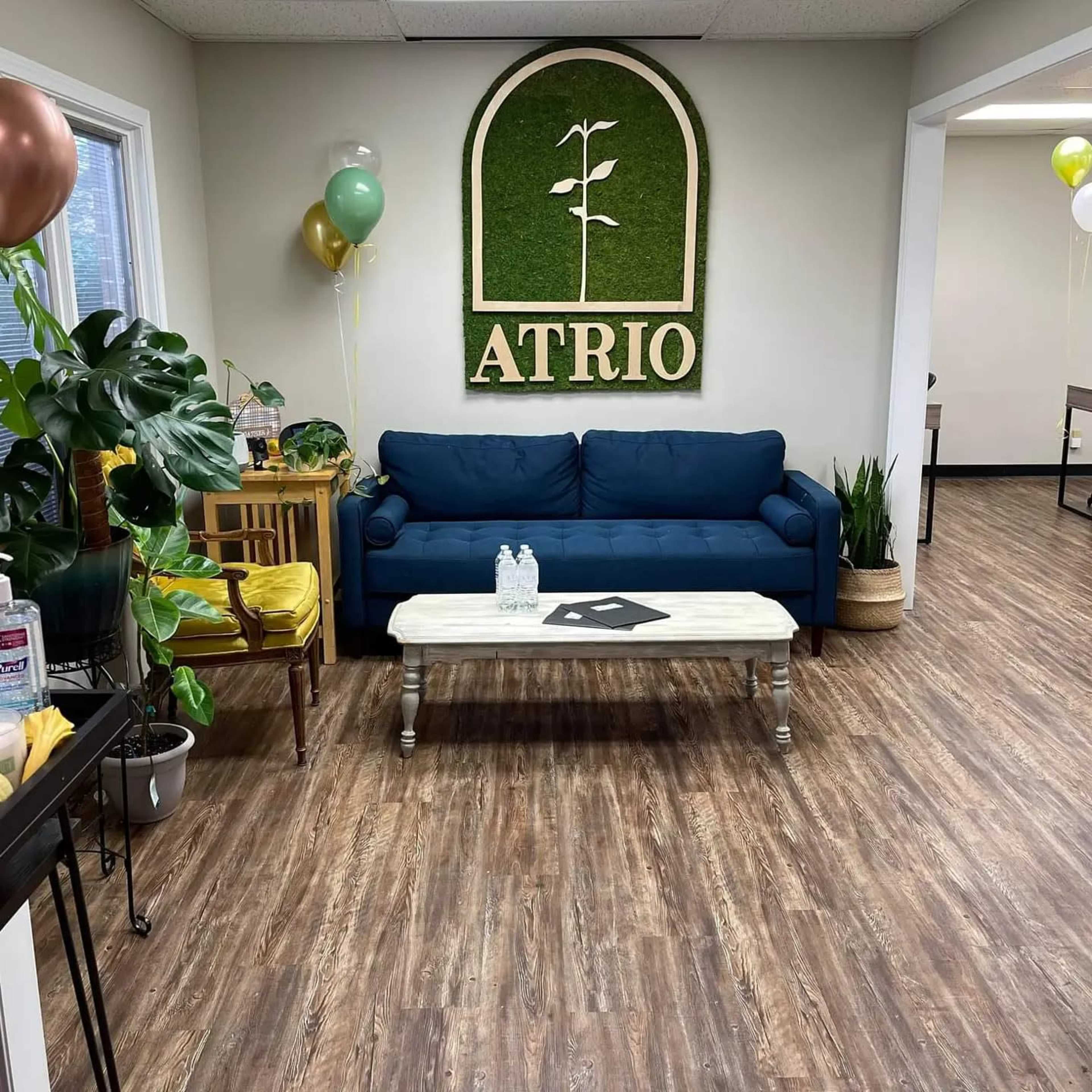 A waiting area with a blue couch, a white coffee table, and a green wall decoration that reads "ATRIO," surrounded by plants and balloons.