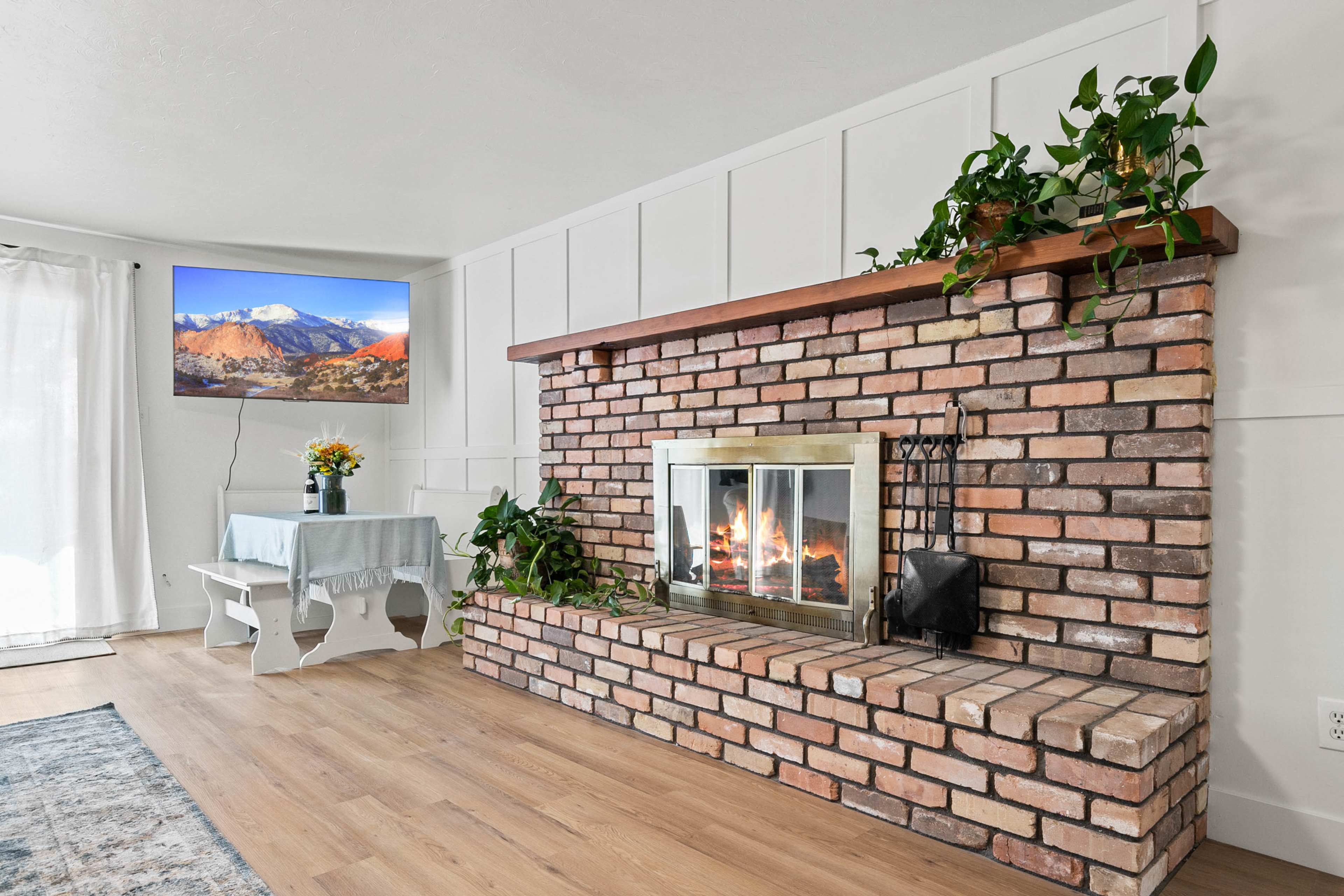 A living room featuring a brick fireplace with a wooden mantle, a small table with flowers, and a television mounted on the wall displaying a mountain scene.
