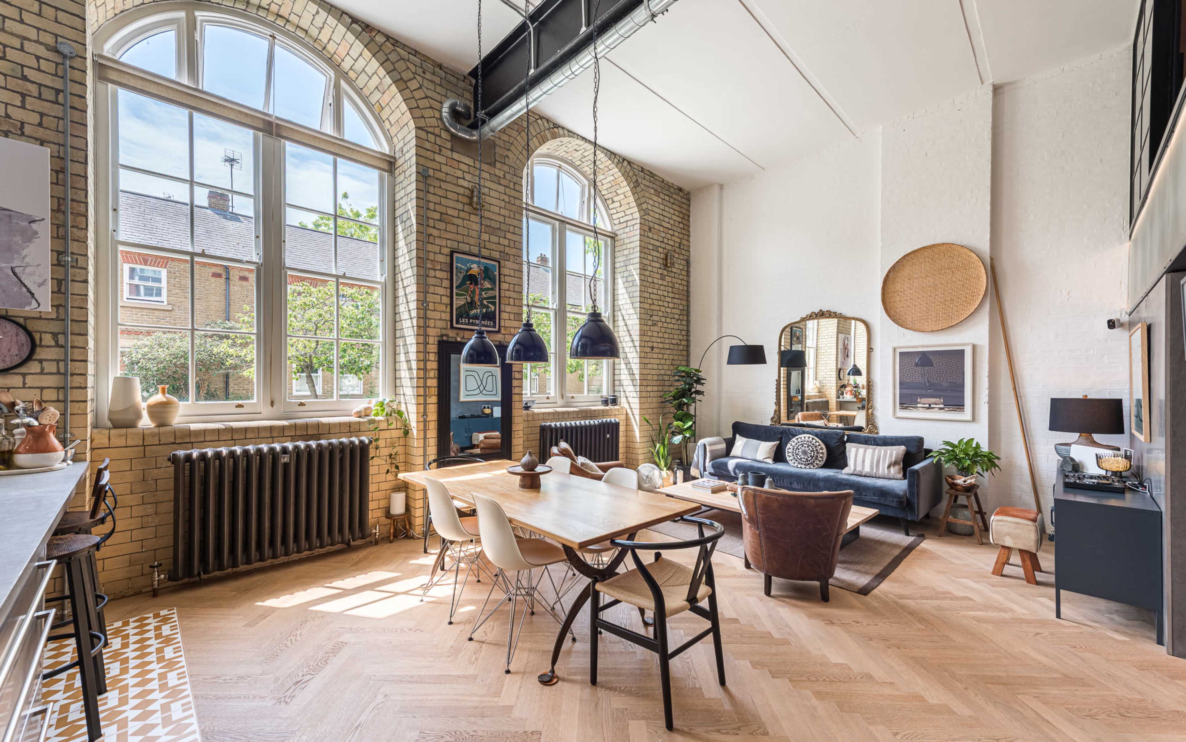 The image shows a spacious living area with tall arched windows, a dining table, and a mix of seating arrangements, set in a room featuring exposed brick walls and wooden flooring.