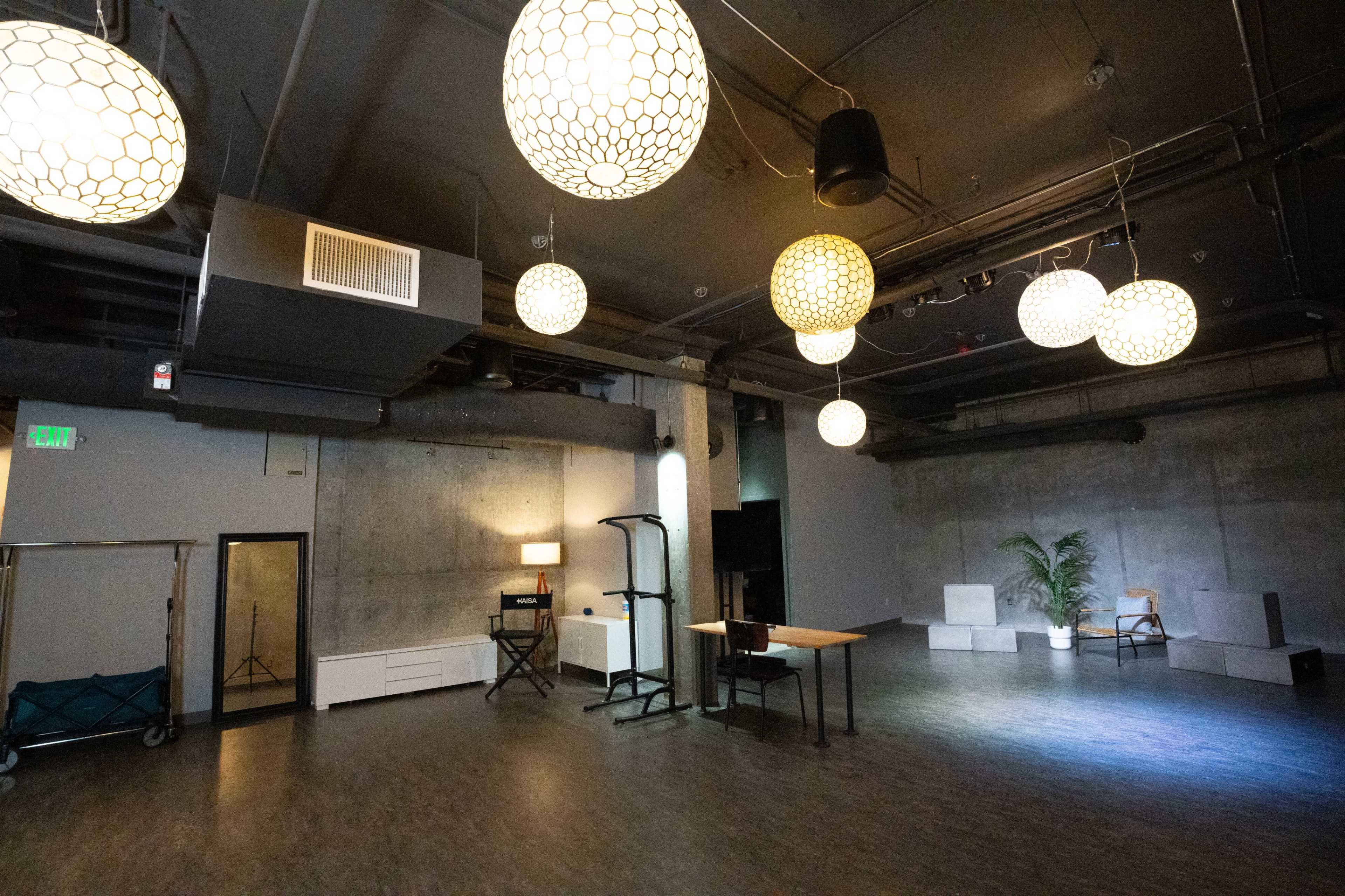 A spacious interior room features several decorative pendant lights, a wooden table, and a plant, with concrete walls and a dark floor.