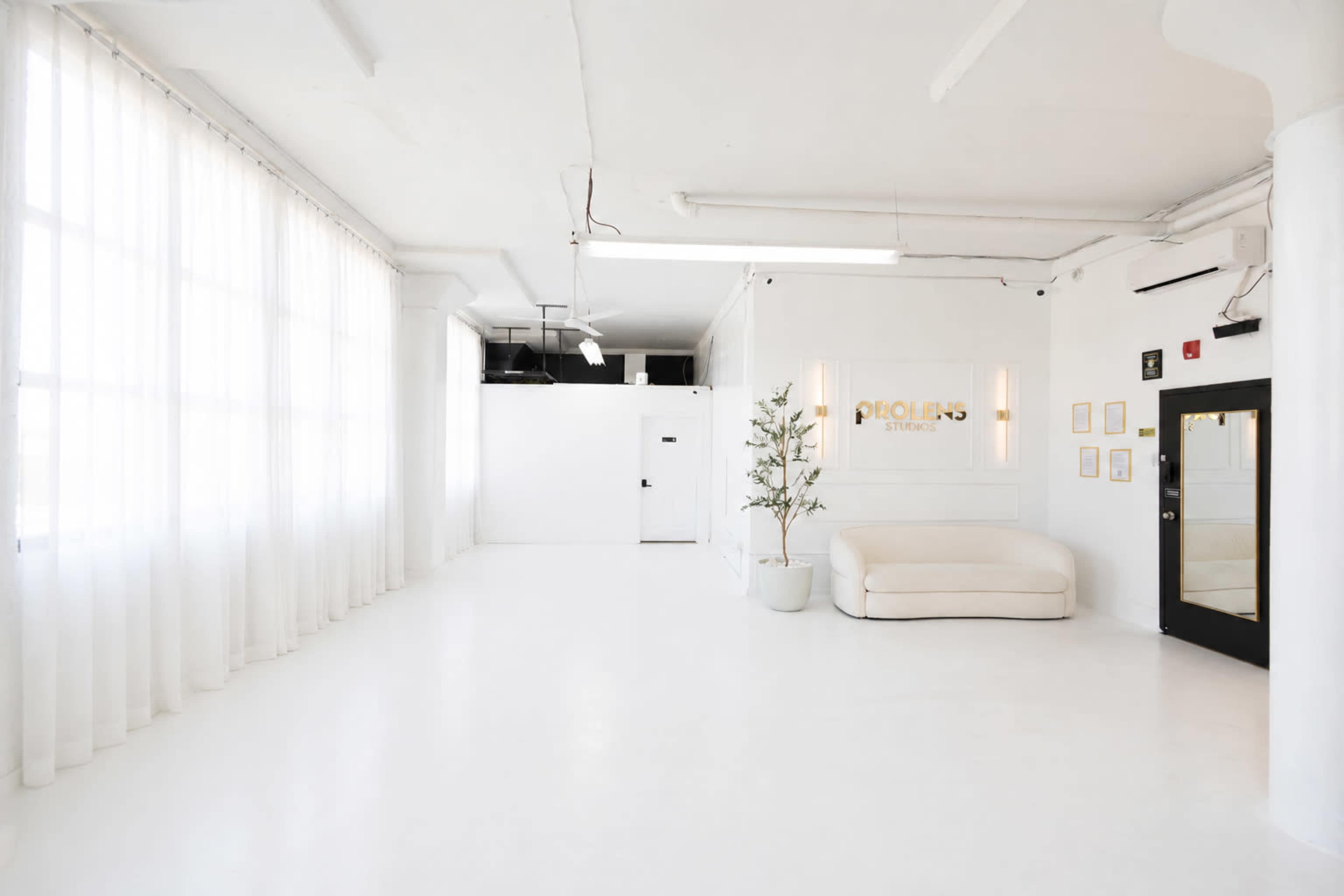 The image shows a spacious, well-lit studio featuring white walls, large windows with sheer curtains, and minimalistic furniture.