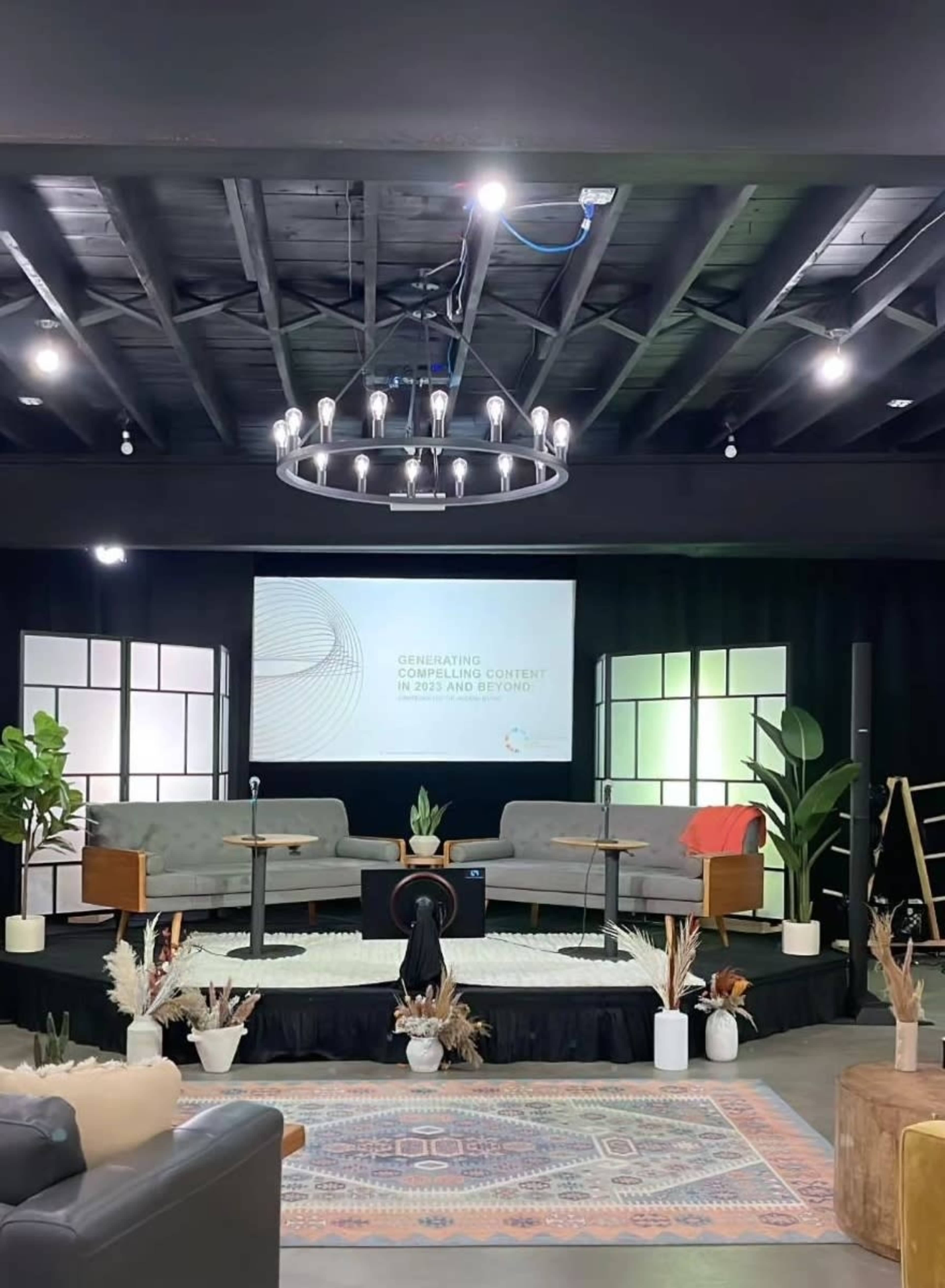 A modern event space features two chairs and a table on a patterned rug, with a large screen displaying text in the background and plants placed around the area.