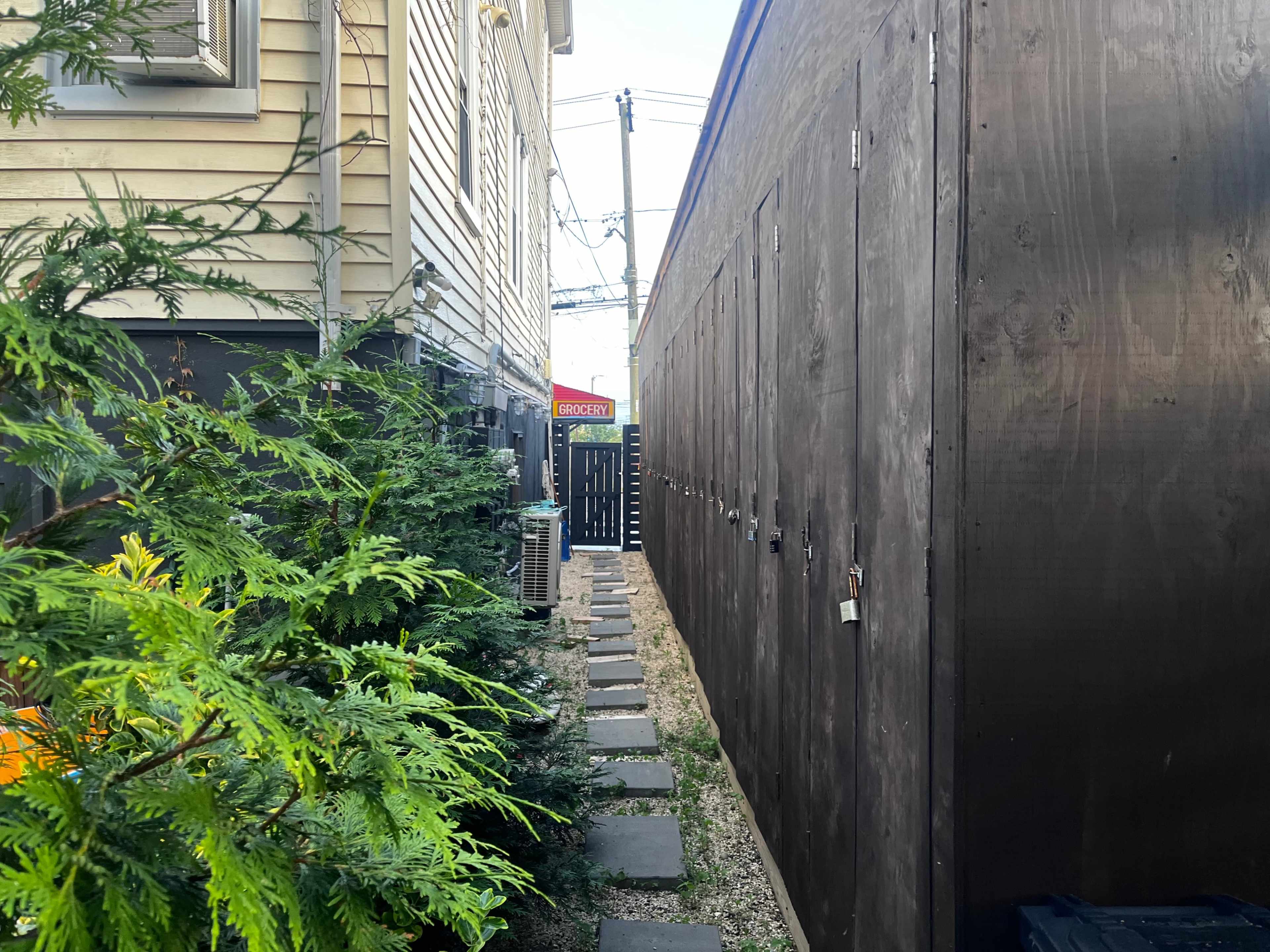 A narrow alleyway lined with wooden structures is flanked by a building and greenery, leading to a visible sign in the distance.