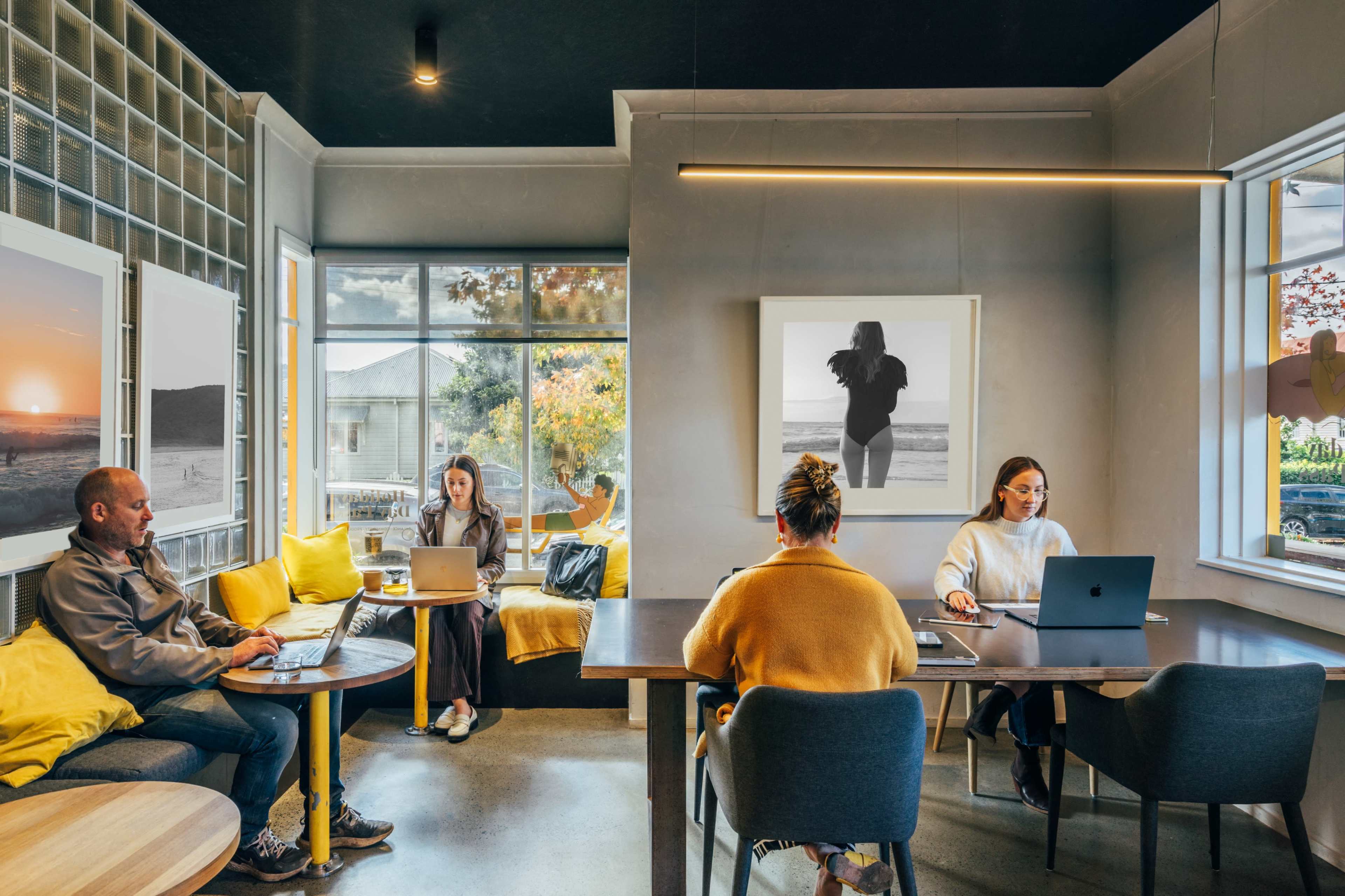 A cozy café interior features several people working on laptops at different tables, surrounded by large windows and artistic wall decor.