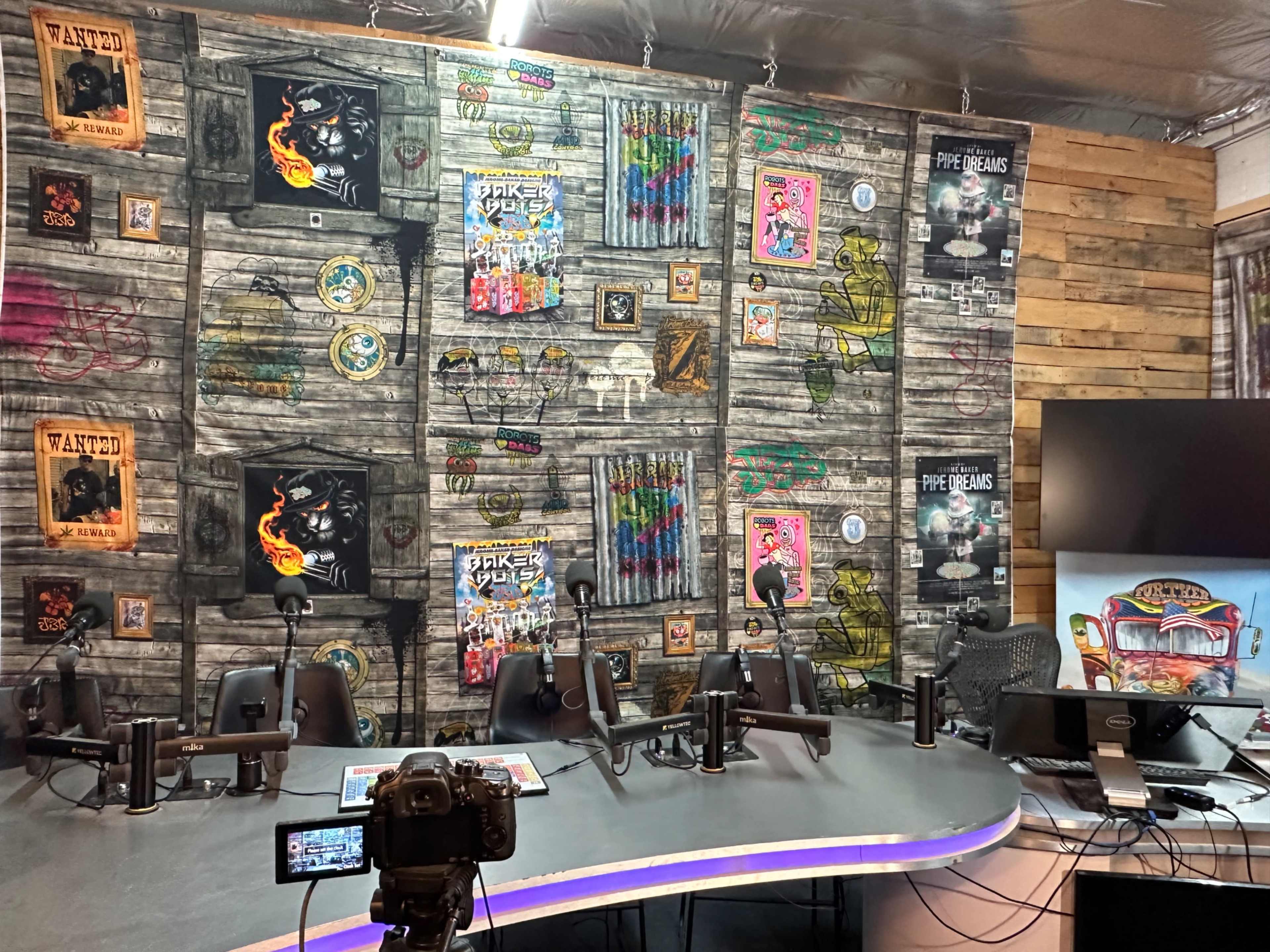 The image shows a podcast recording studio with a large, curved table, multiple microphones, and a colorful, patterned wall featuring various posters and artwork.