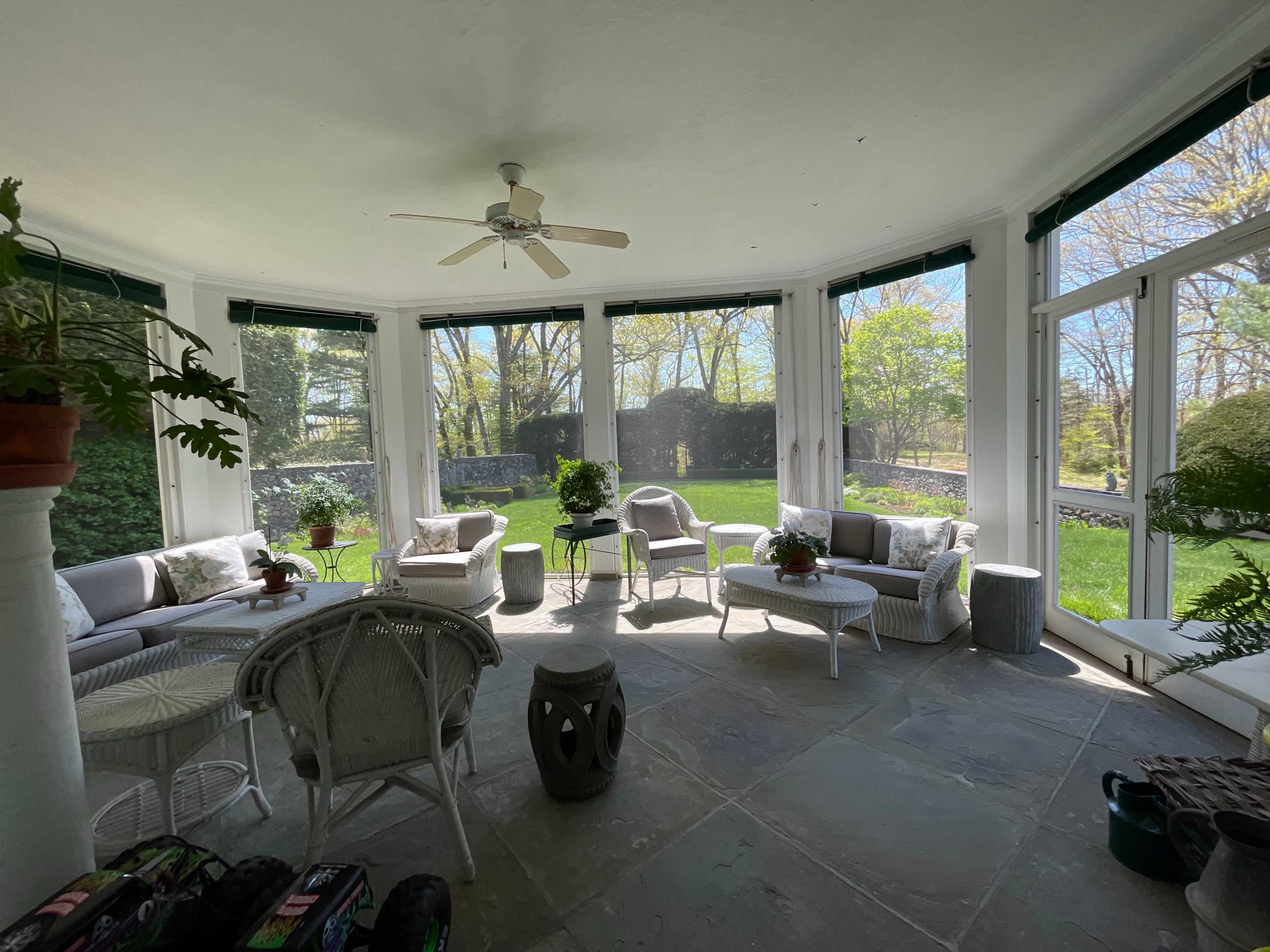 The image shows a sunlit sunroom with multiple seating areas and large windows overlooking a green outdoor space.
