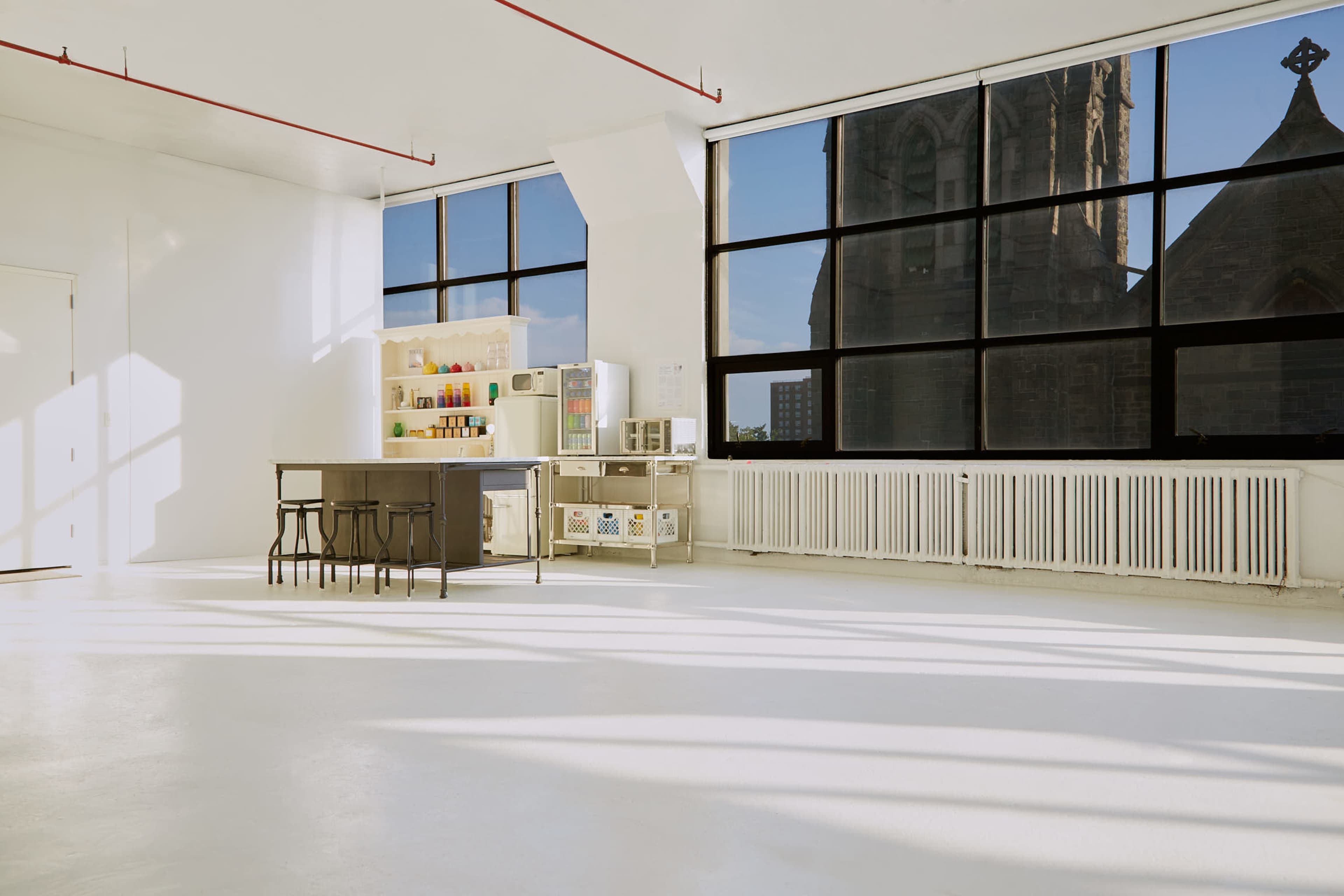 A bright, empty room features a kitchen island and a shelf of colorful jars, with large windows overlooking a stone church.