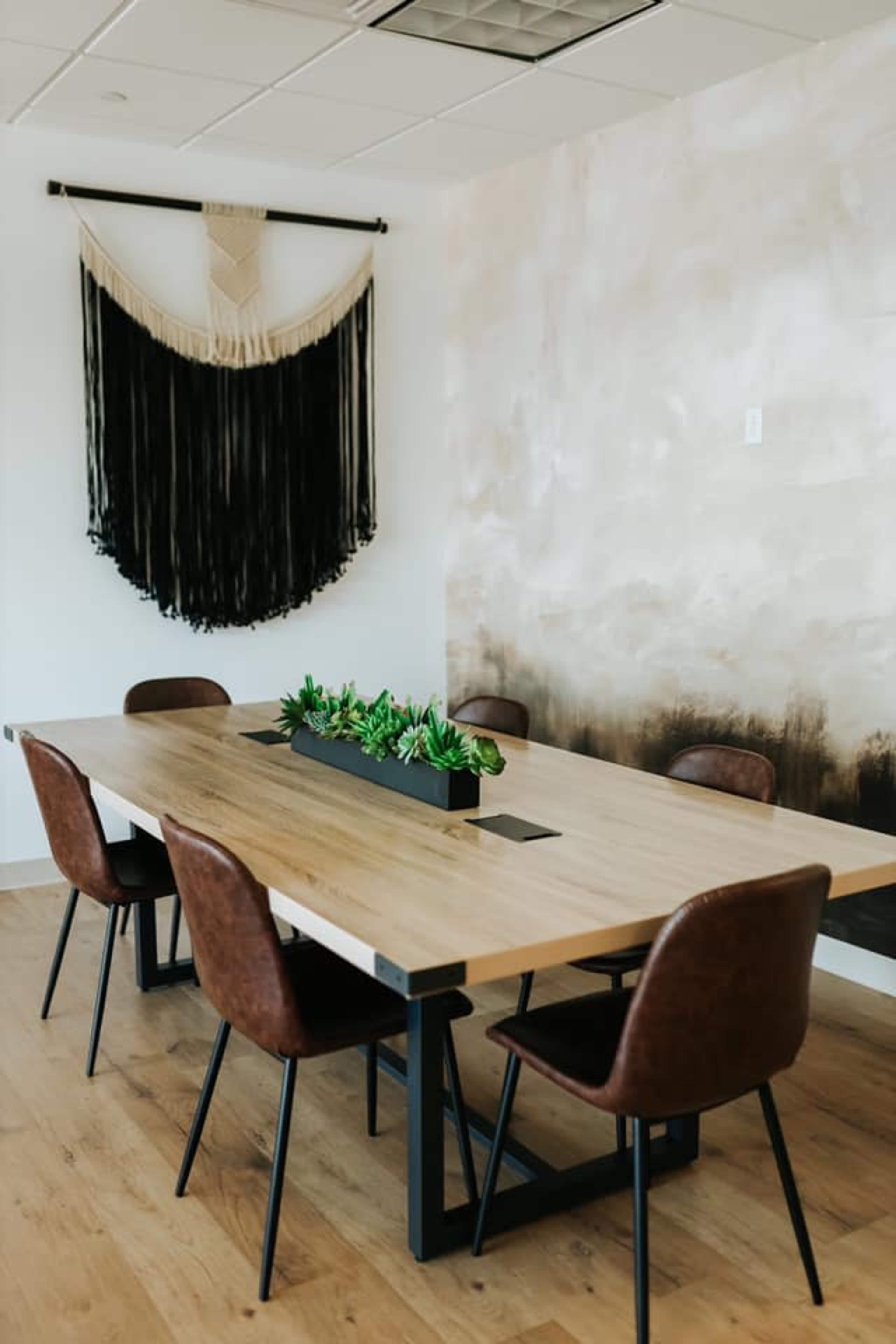 A long wooden table with six brown chairs is positioned in a modern meeting room, decorated with a textured wall and a wall hanging made of fabric.