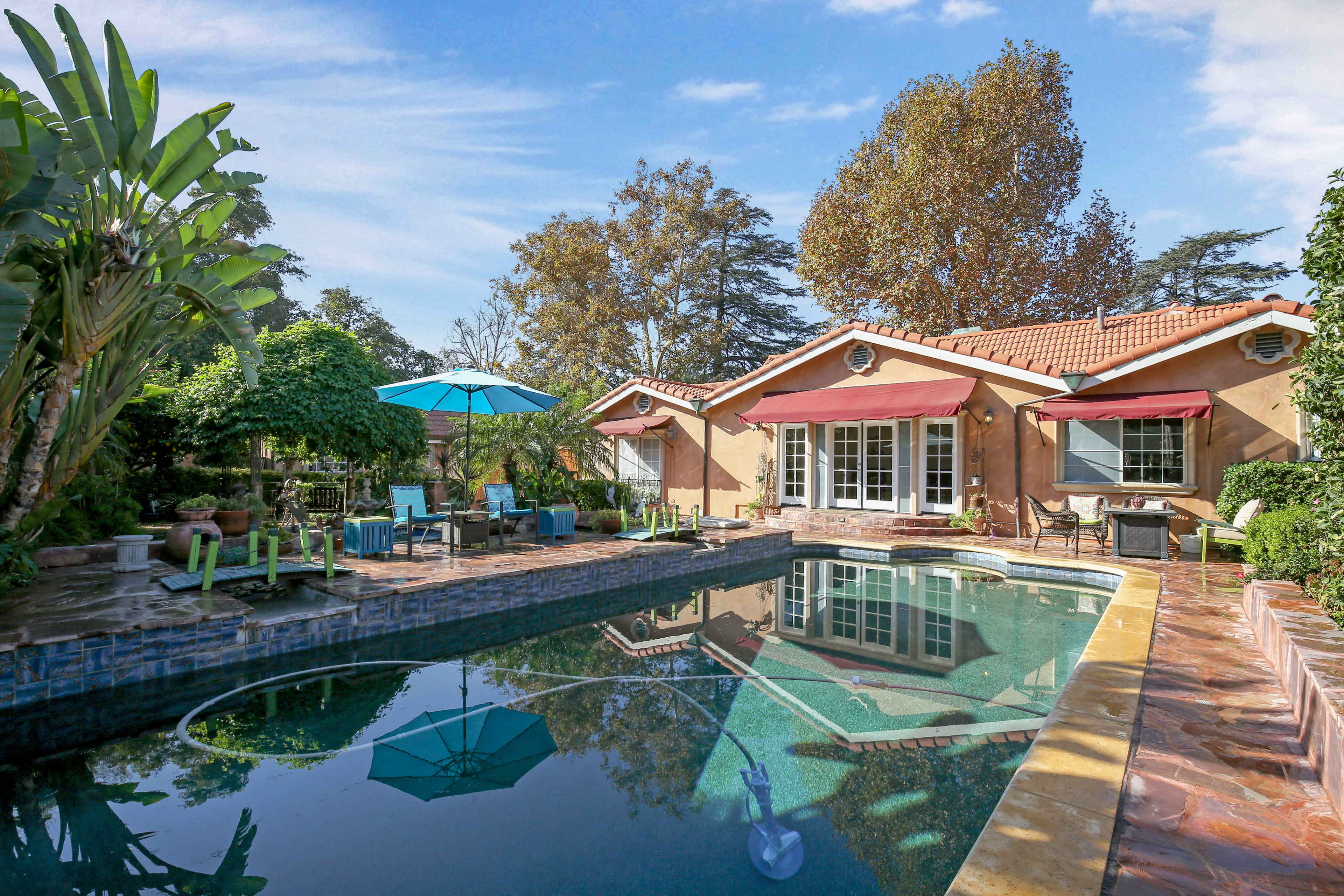 The image shows a backyard with a swimming pool surrounded by lounge chairs and an umbrella, adjacent to a house with a landscaped garden.