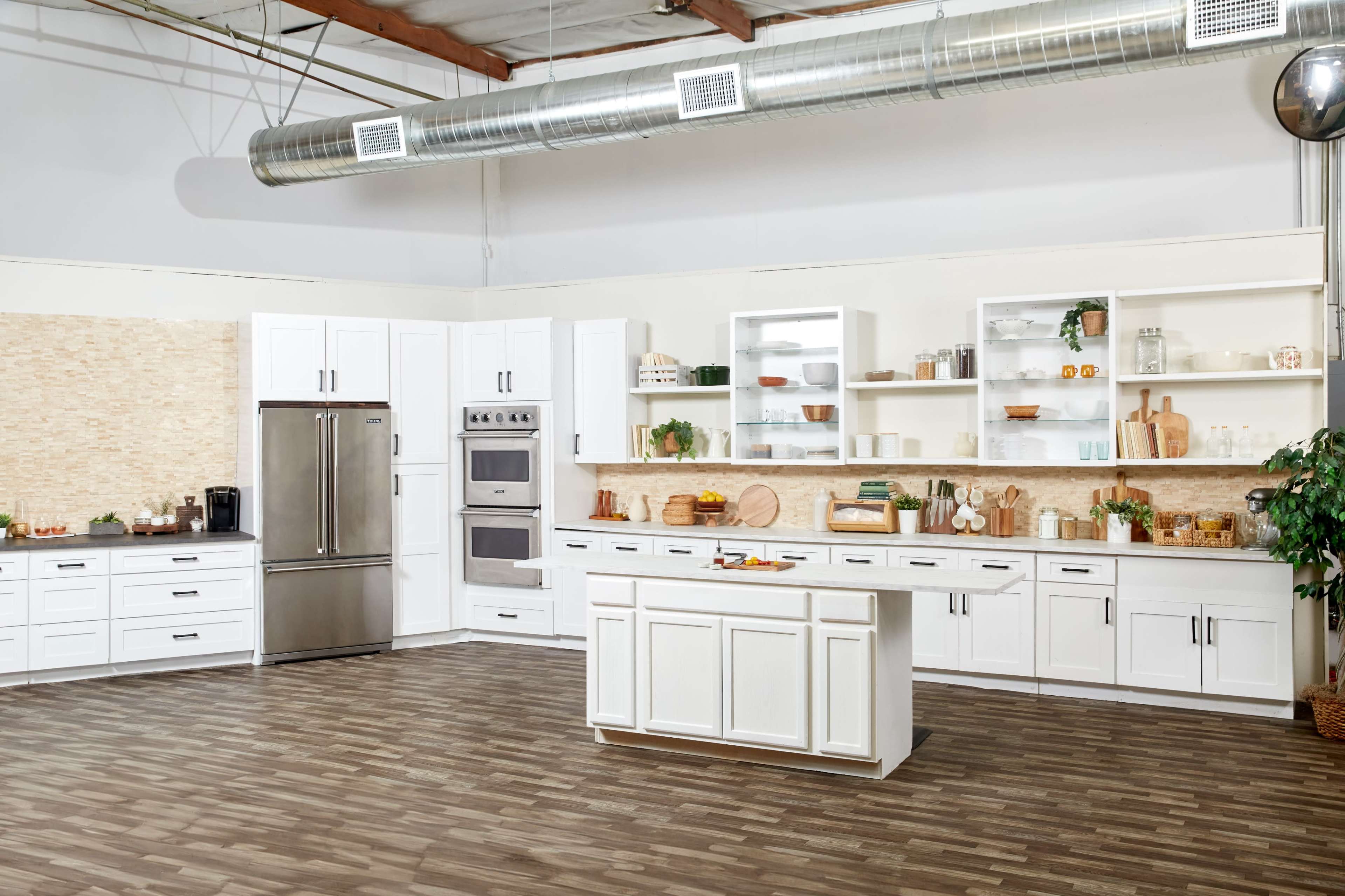 A modern kitchen with white cabinetry, stainless steel appliances, and an island, featuring a light wood floor and various kitchen items displayed on shelves.