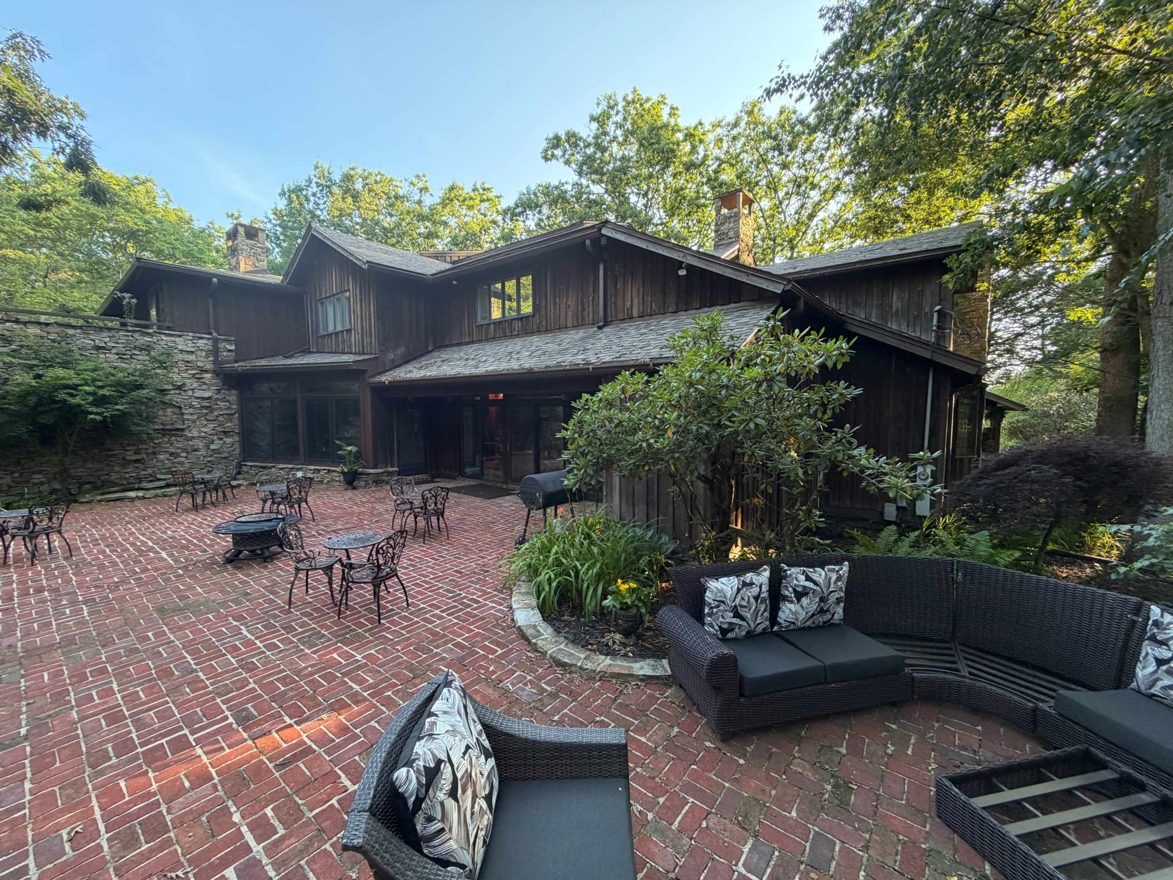 A rustic wooden house surrounded by a brick patio, with several outdoor seating areas and lush greenery.