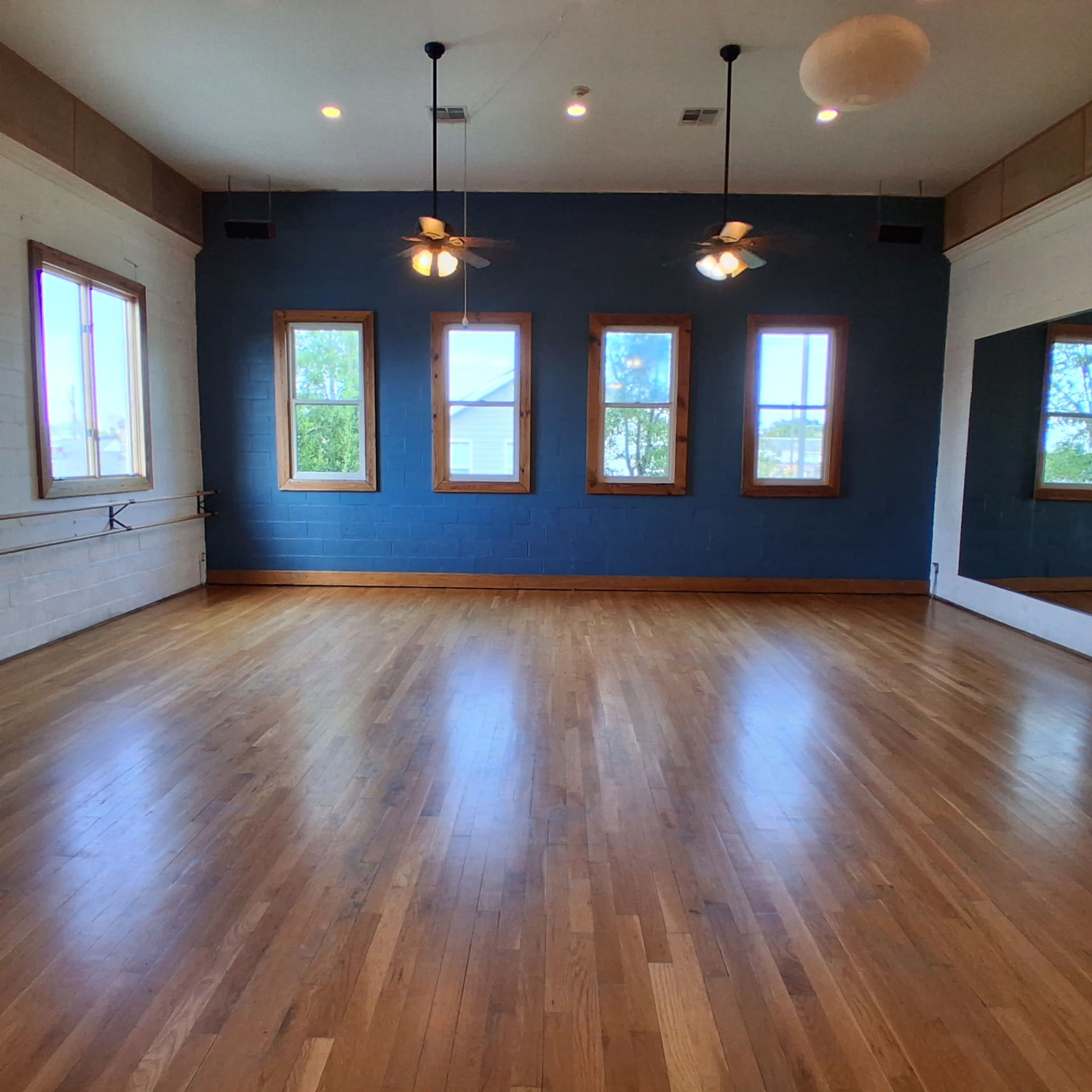 The image shows a spacious, empty room with wooden flooring, blue walls, large windows, and ceiling fans.