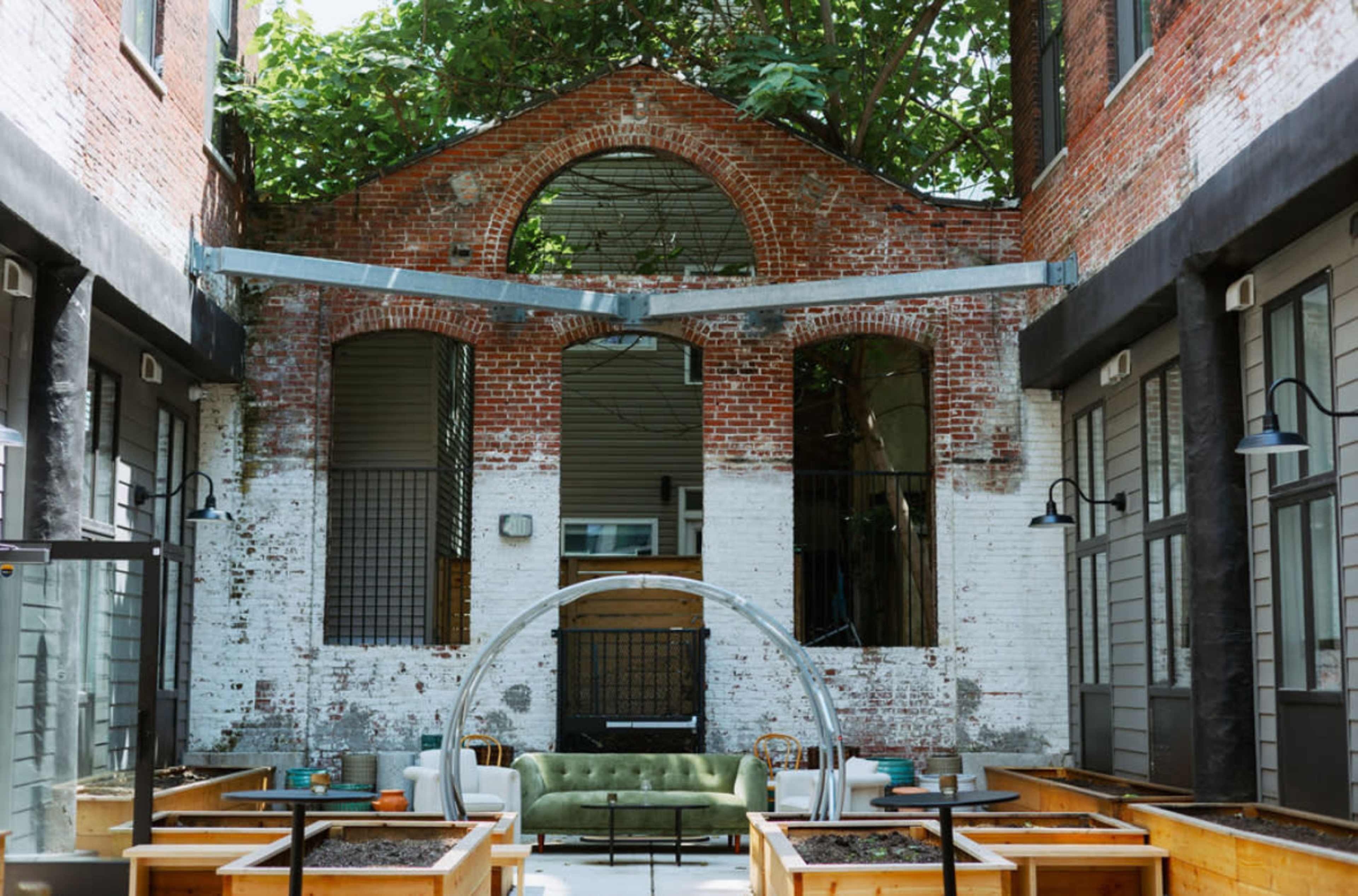 The image shows an open courtyard with exposed brick walls, large windows, and a green sofa surrounded by wooden planters.