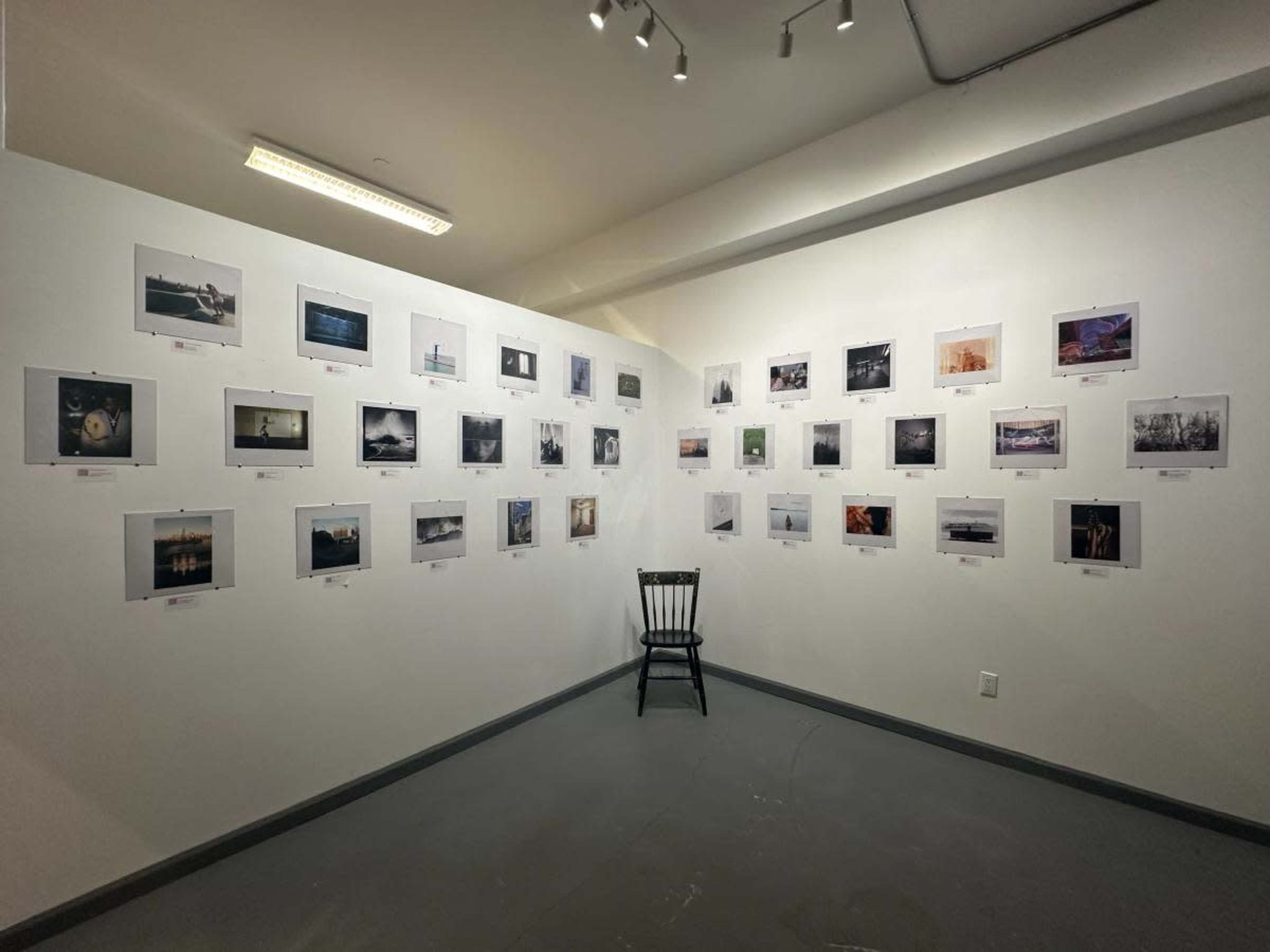 A gallery space features a wall display of various photographs, with a single wooden chair positioned in the center.
