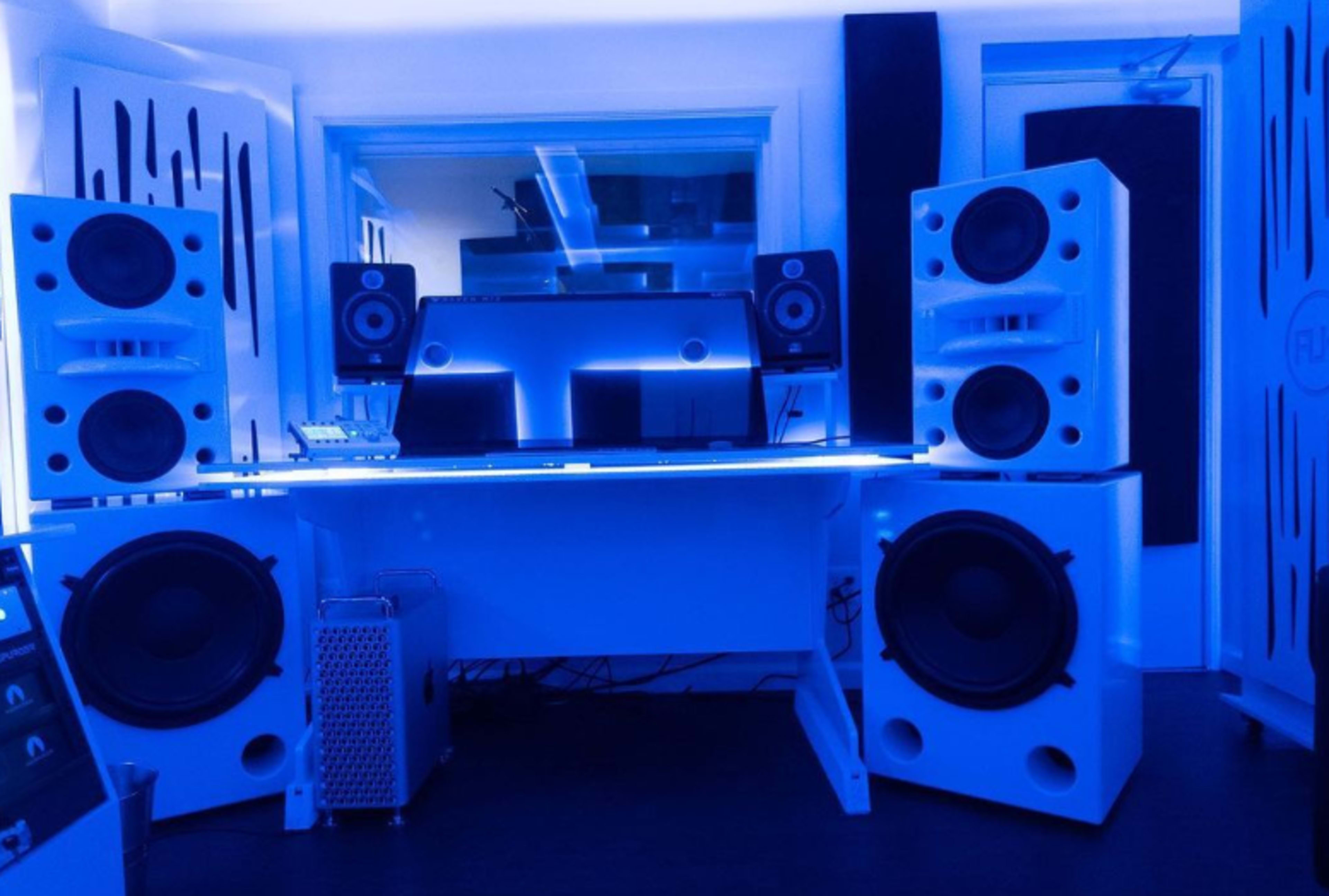 A modern music studio with blue lighting, featuring a central desk, large speakers on either side, and studio monitors positioned on top.