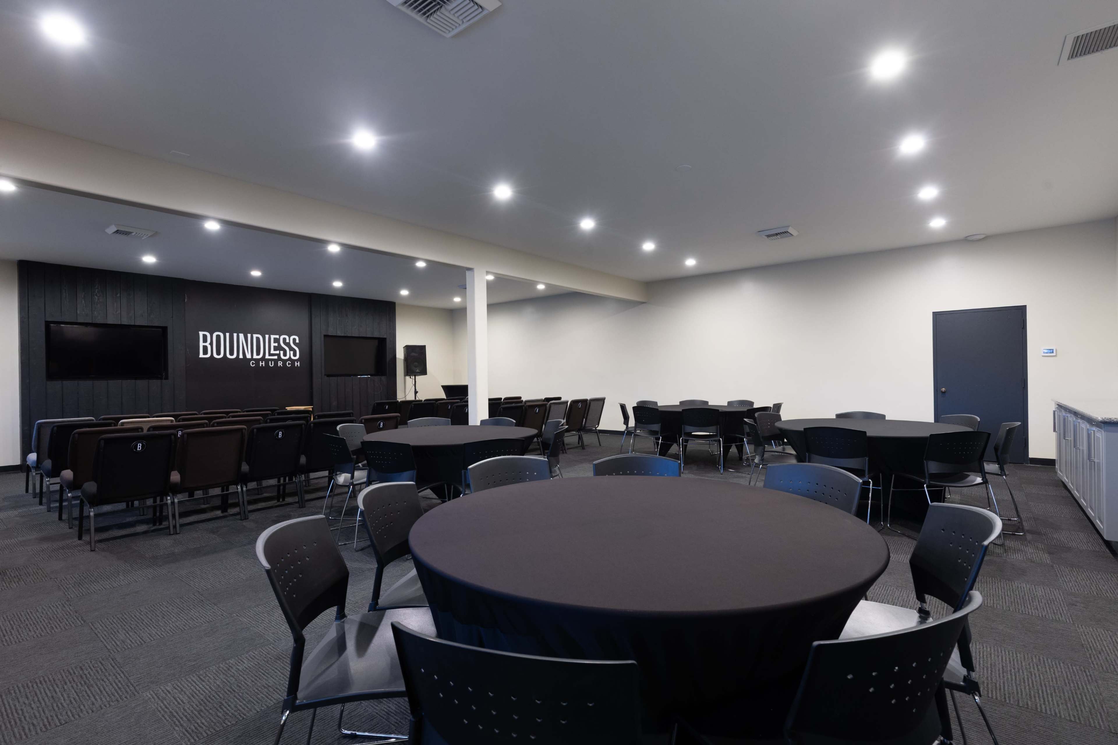The image shows an indoor event space with round tables, chairs, and a presentation area featuring a screen and a sign that reads "BOUNDLESS."