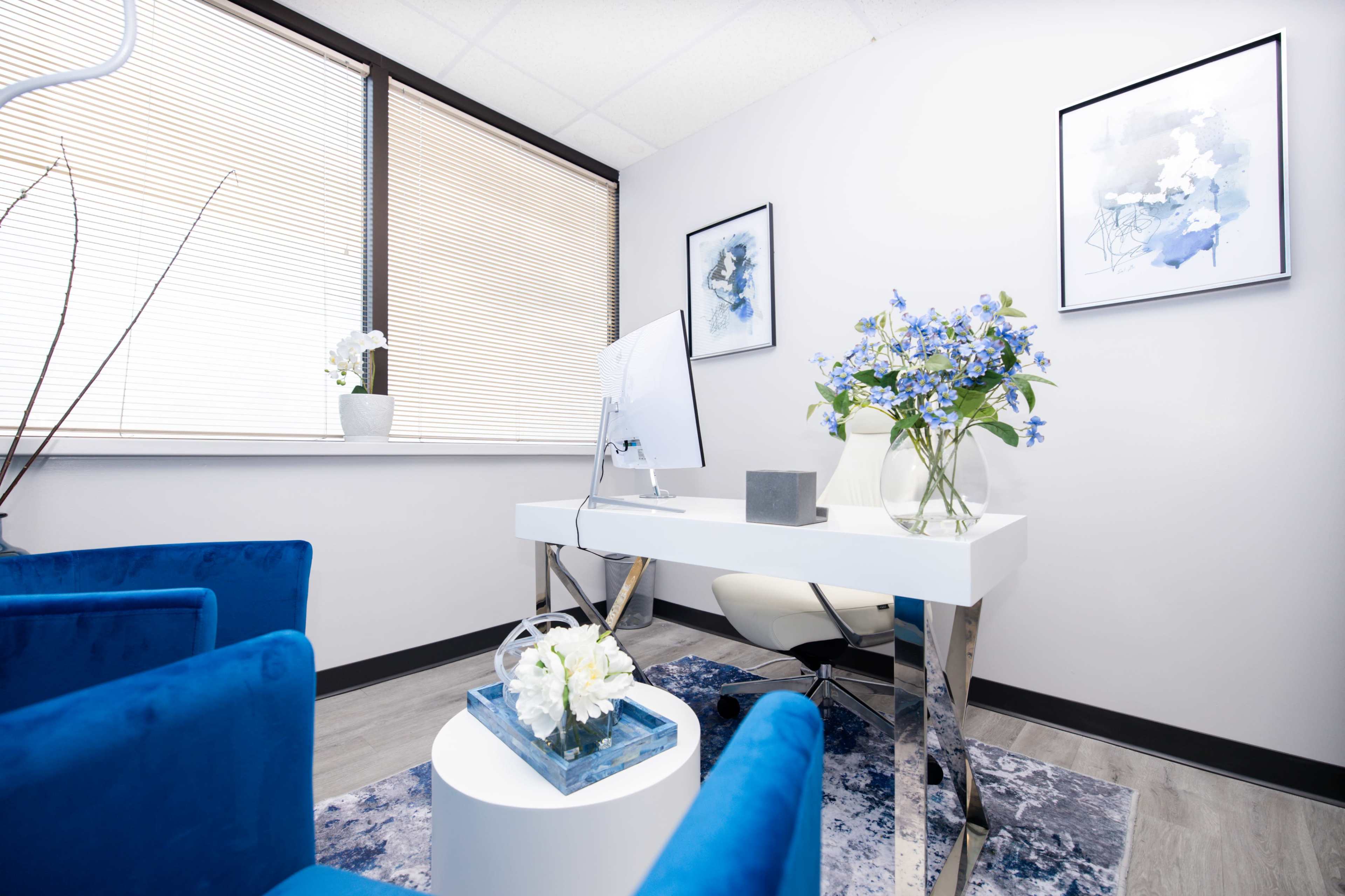 A modern office features a white desk with a computer, a blue floral arrangement, and blue chairs, all set against a backdrop of large windows with blinds.