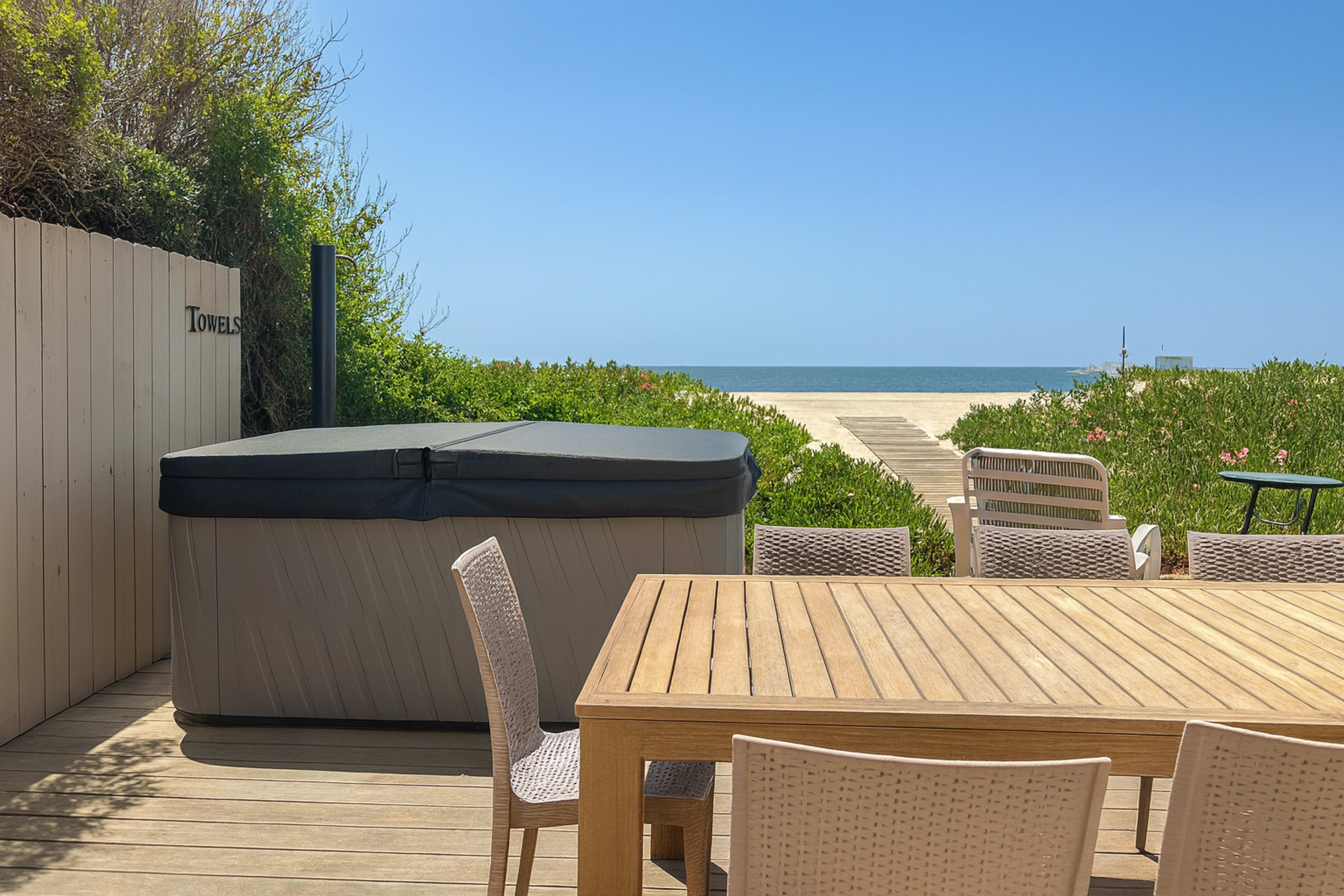 A wooden dining table with chairs is set on a deck beside a hot tub, overlooking a sandy beach and the ocean.