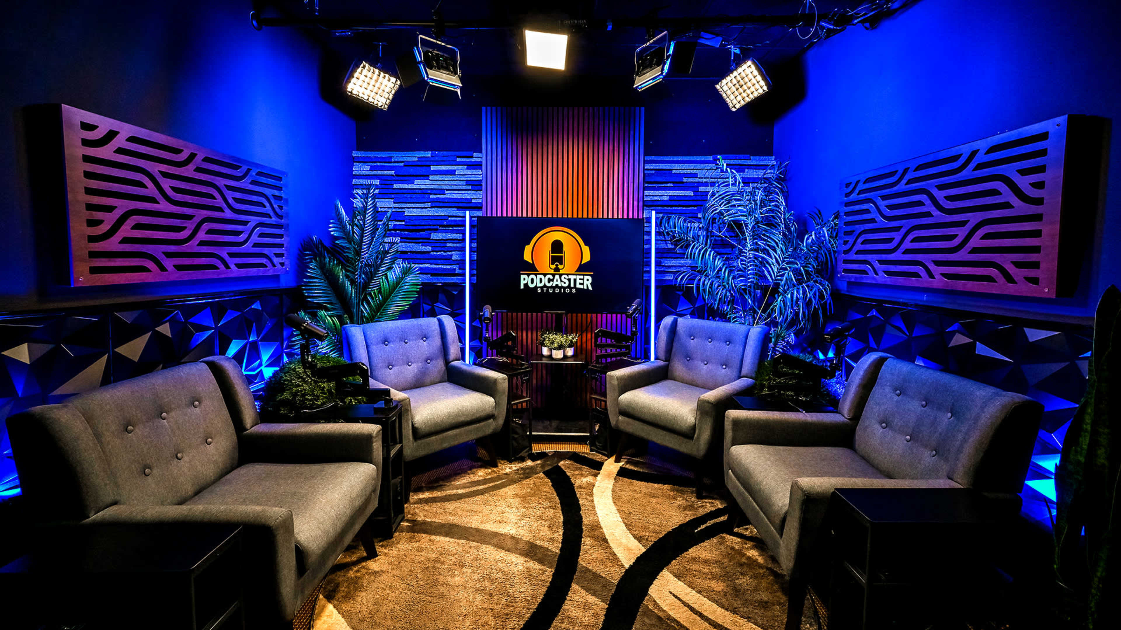 A podcast studio is set up with gray sofas, decorative plants, and a central screen displaying the word "PODCASTER," all illuminated by overhead lights.