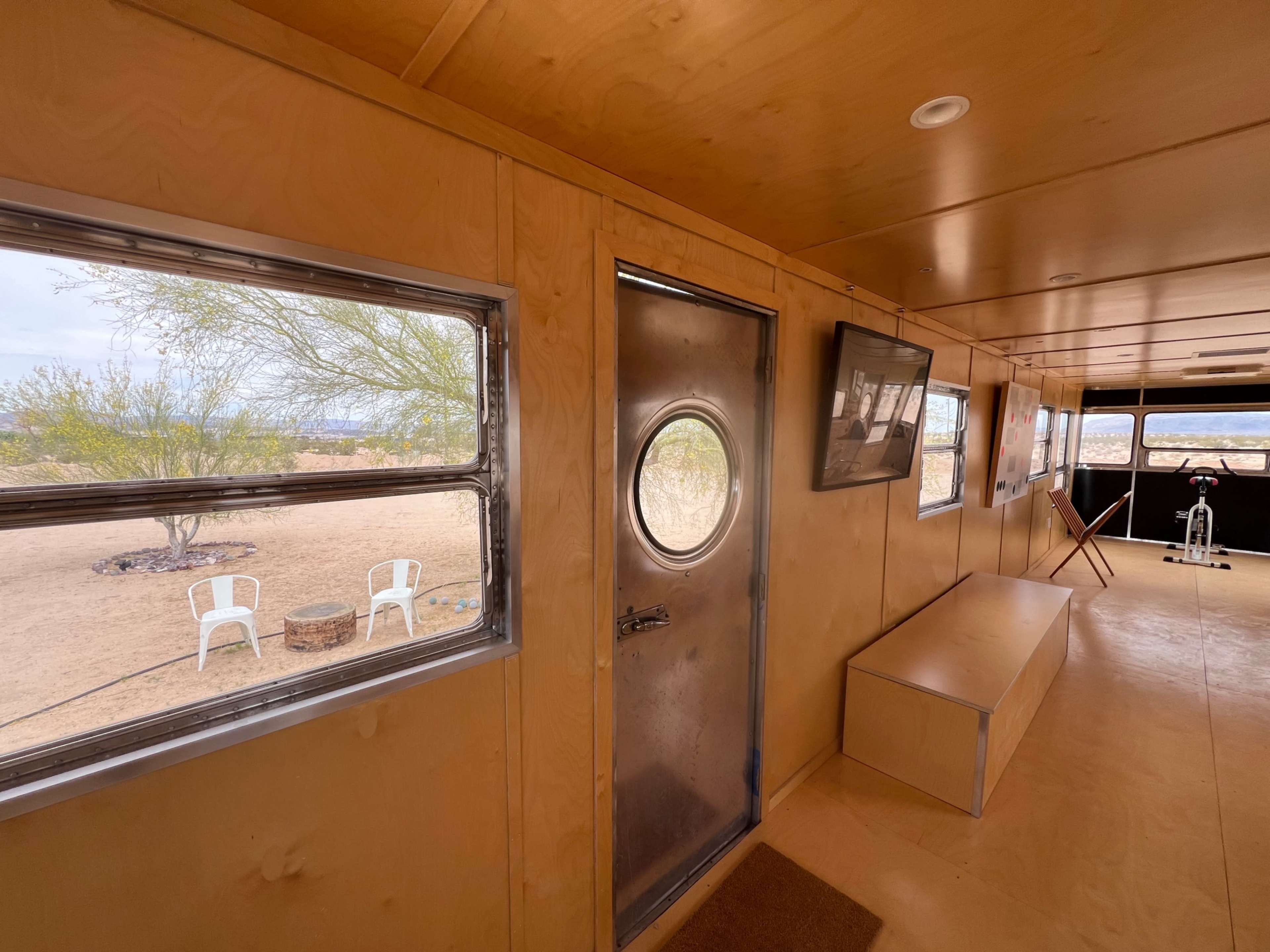 The interior of a small wooden building features a round metal door, a television mounted on a wall, and two white chairs visible through a window.