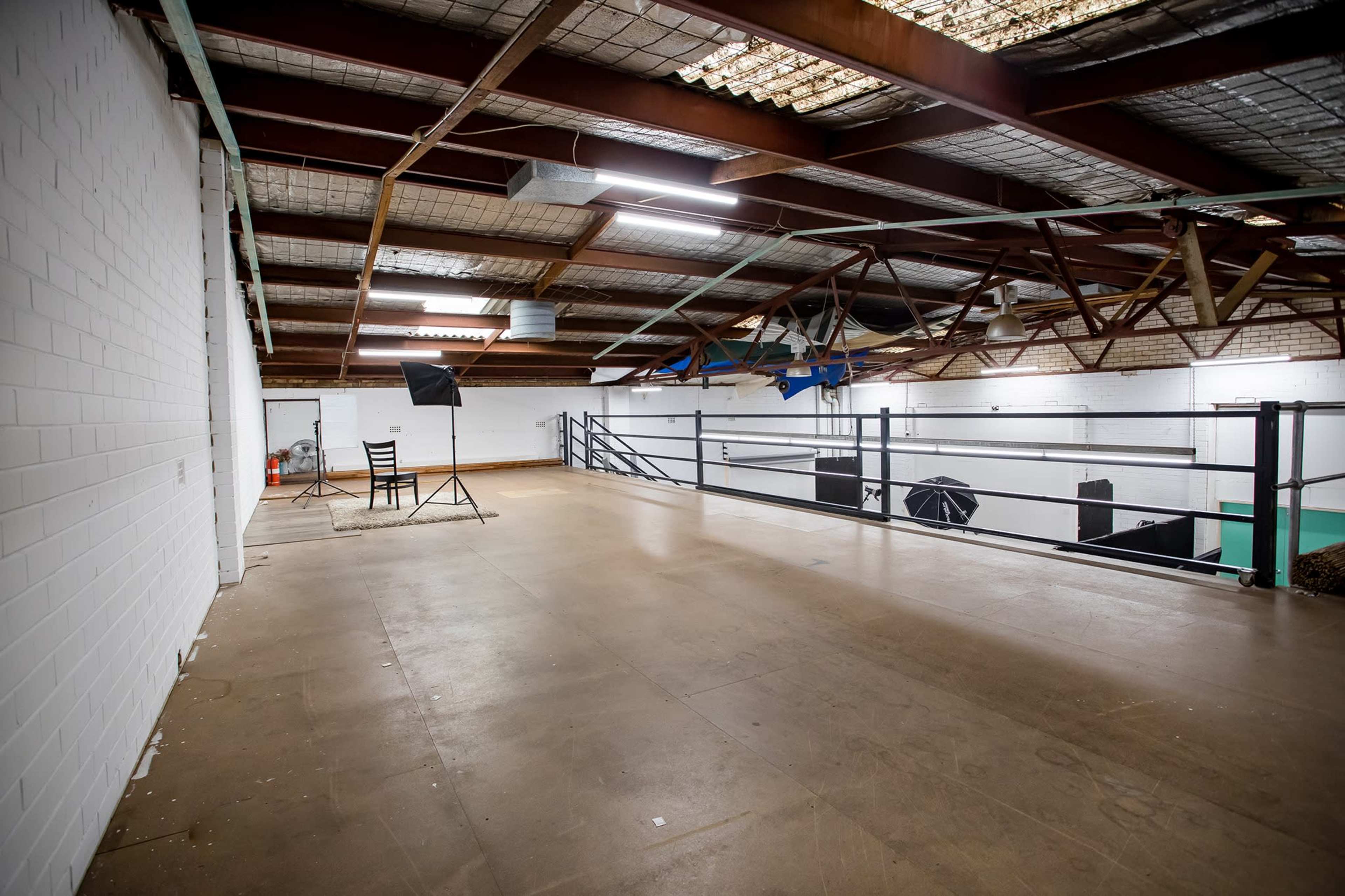A large, empty indoor space with high ceilings, metal beams, and a wooden floor, featuring a chair and lighting equipment set up for a photo shoot.