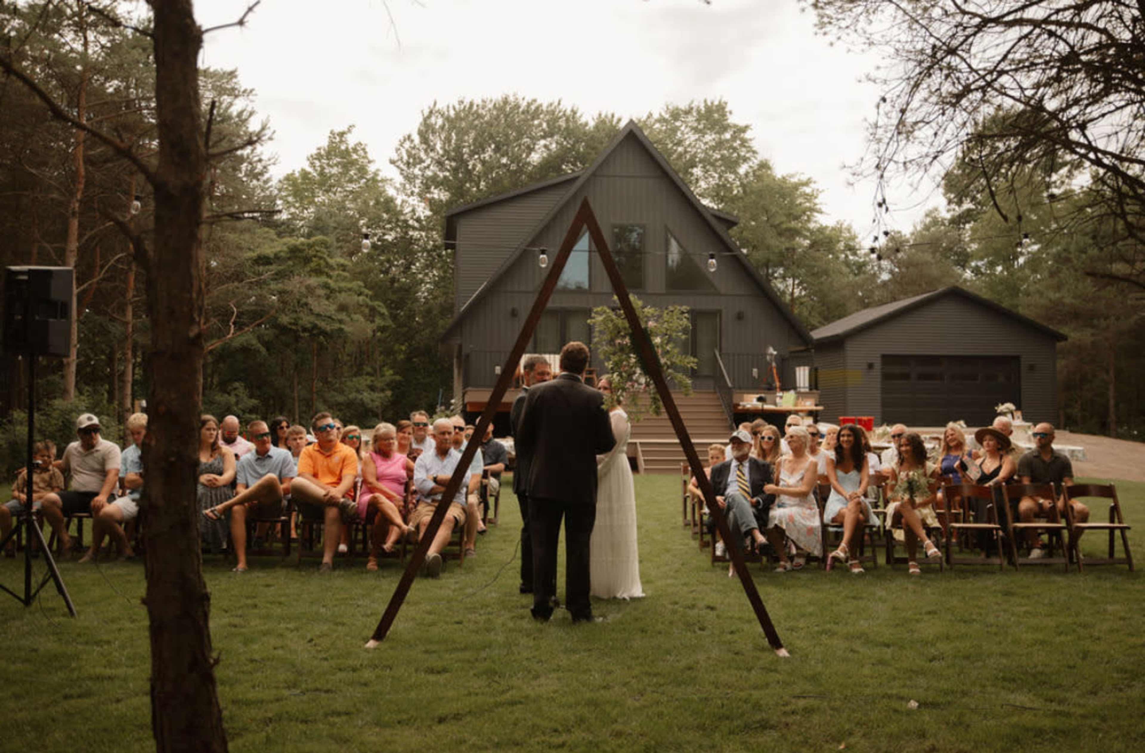 A couple stands under an A-frame structure while guests sit on chairs in a grassy area, with a modern black house in the background.
