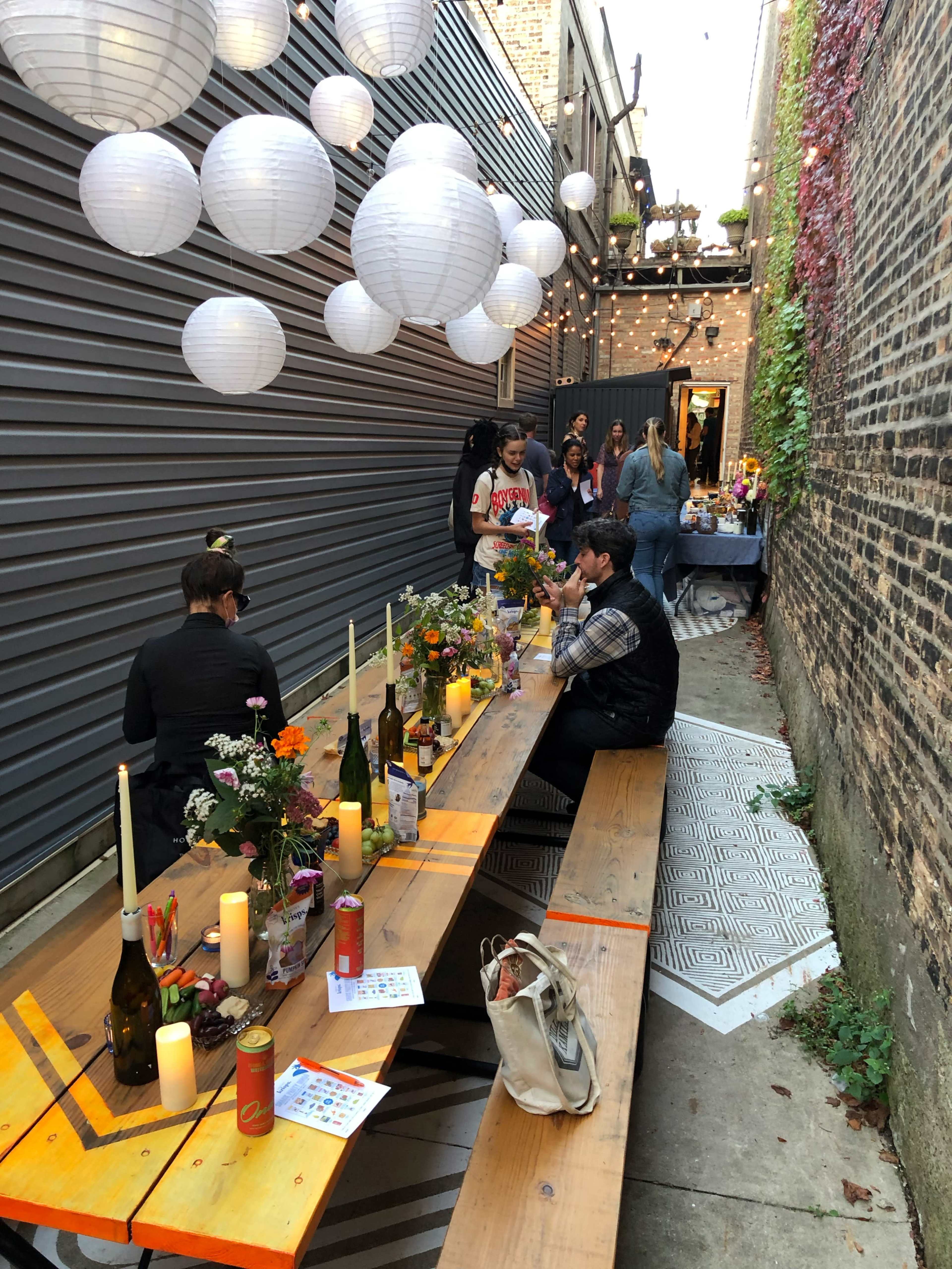 A narrow alley decorated with string lights and paper lanterns, featuring a long wooden table set for a gathering with candles and flowers.