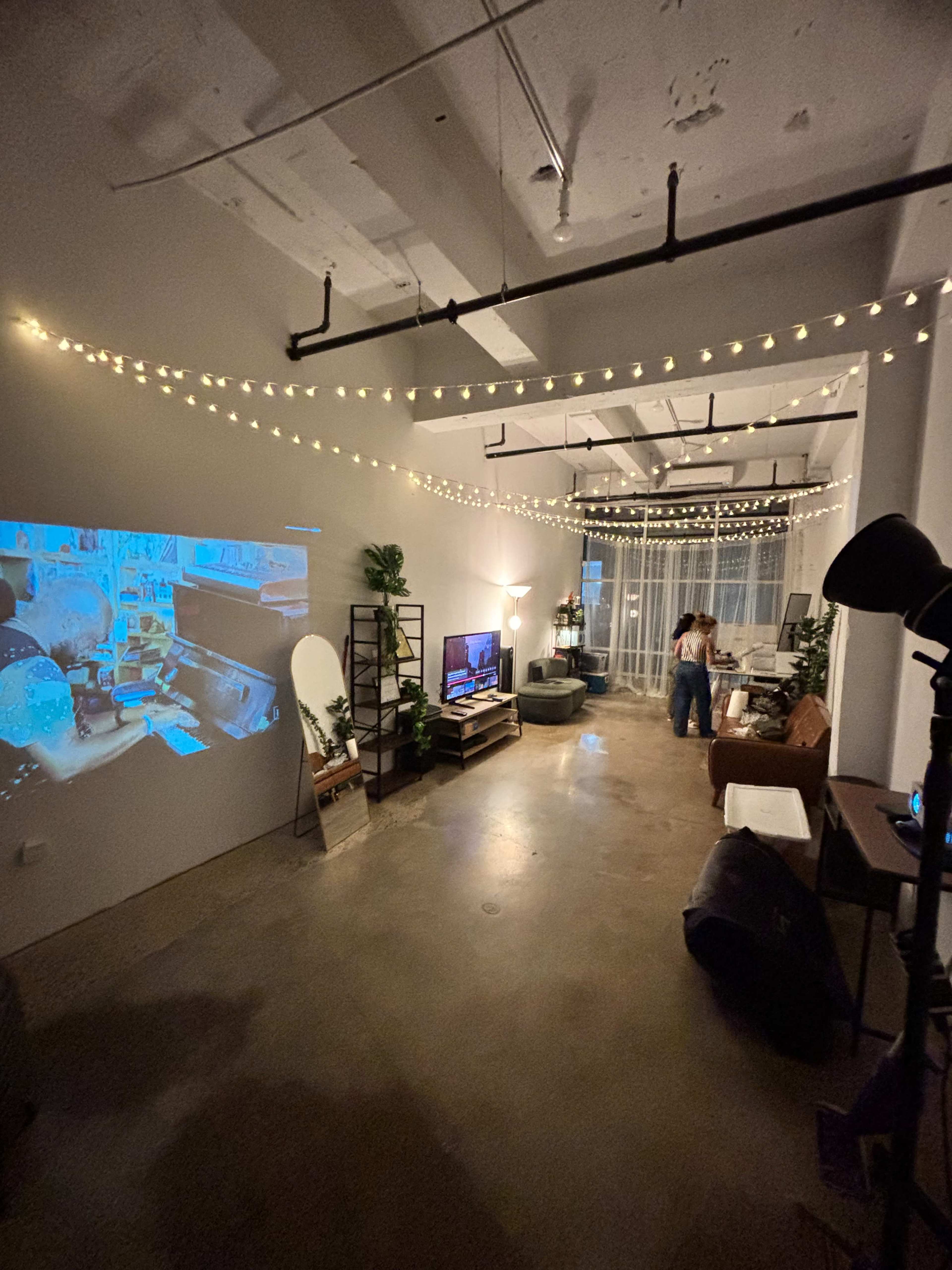 The image shows a modern living space with a projection on the wall, string lights overhead, and various seating areas, including a couch and desk.