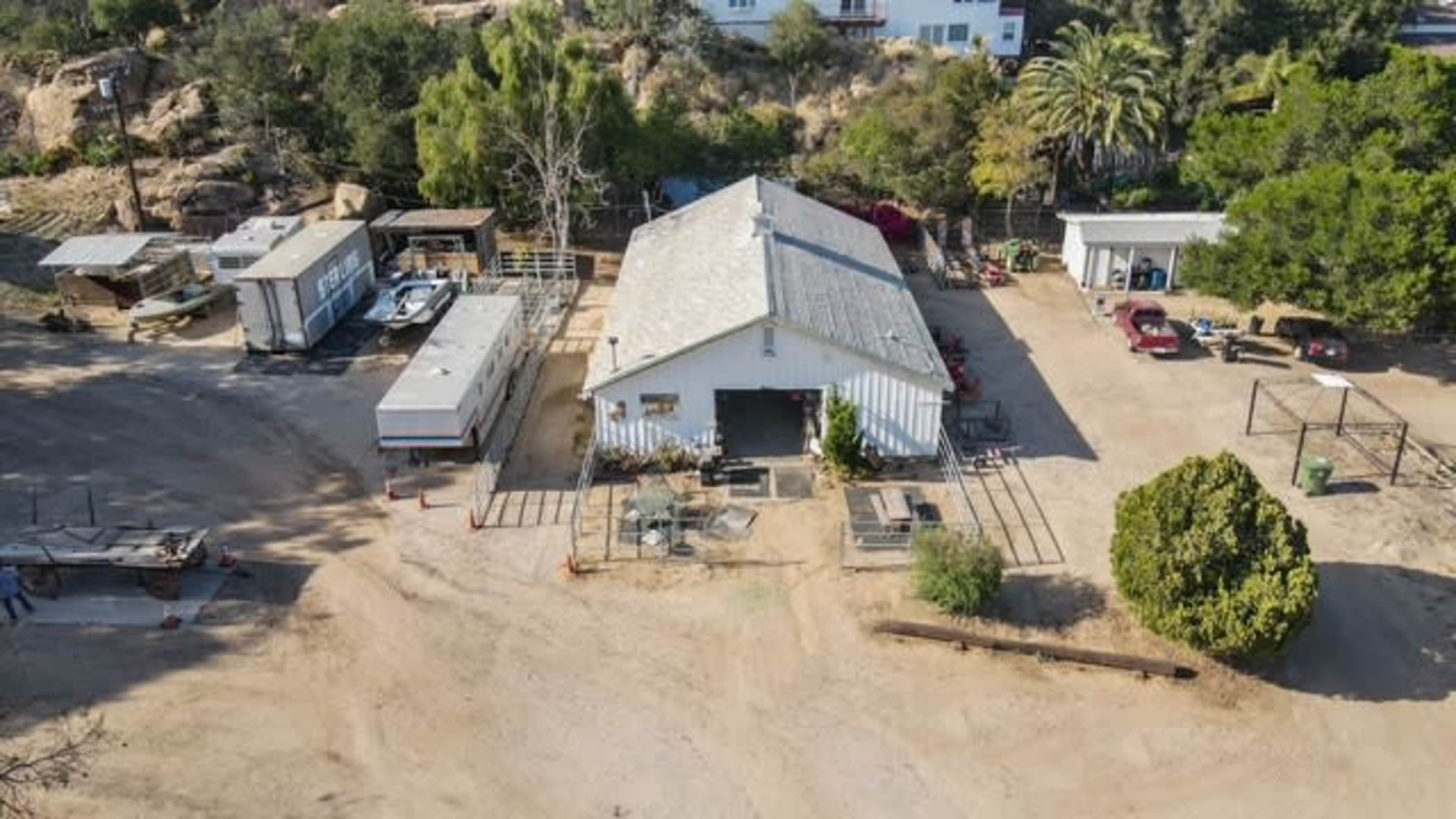 The image shows a large agricultural building surrounded by several smaller structures and vehicles on a sandy lot, with trees and hills in the background.